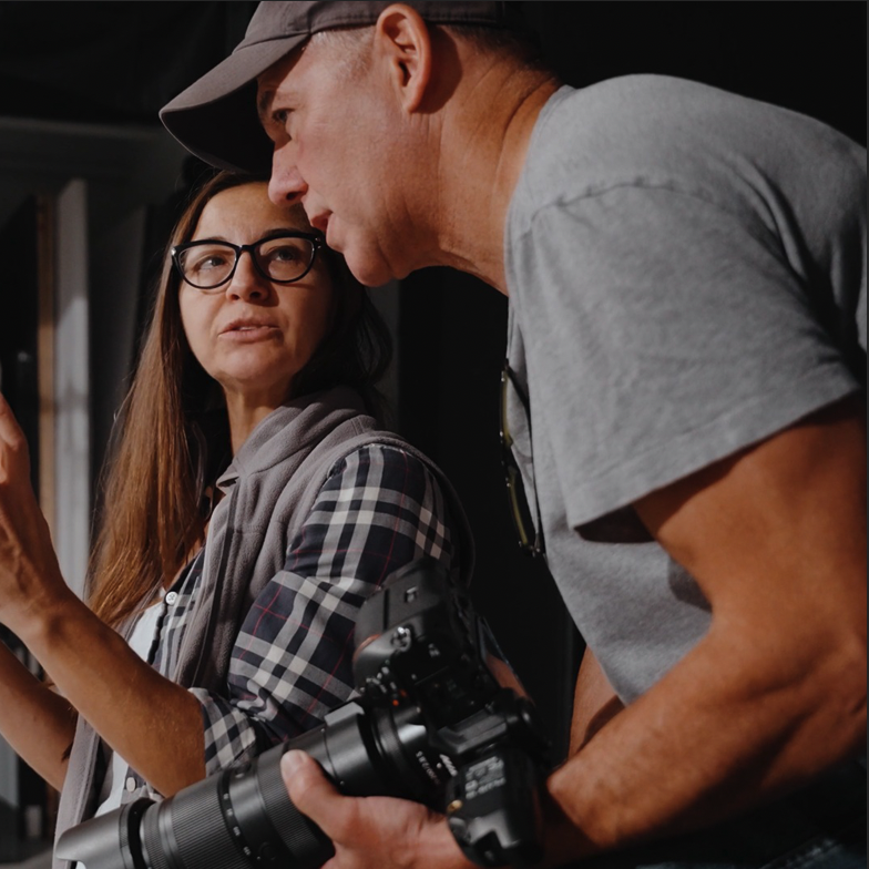 A man and a woman engaged in a conversation; the woman is wearing glasses and a plaid shirt, while the man is holding a camera and wearing a gray t-shirt and a cap.