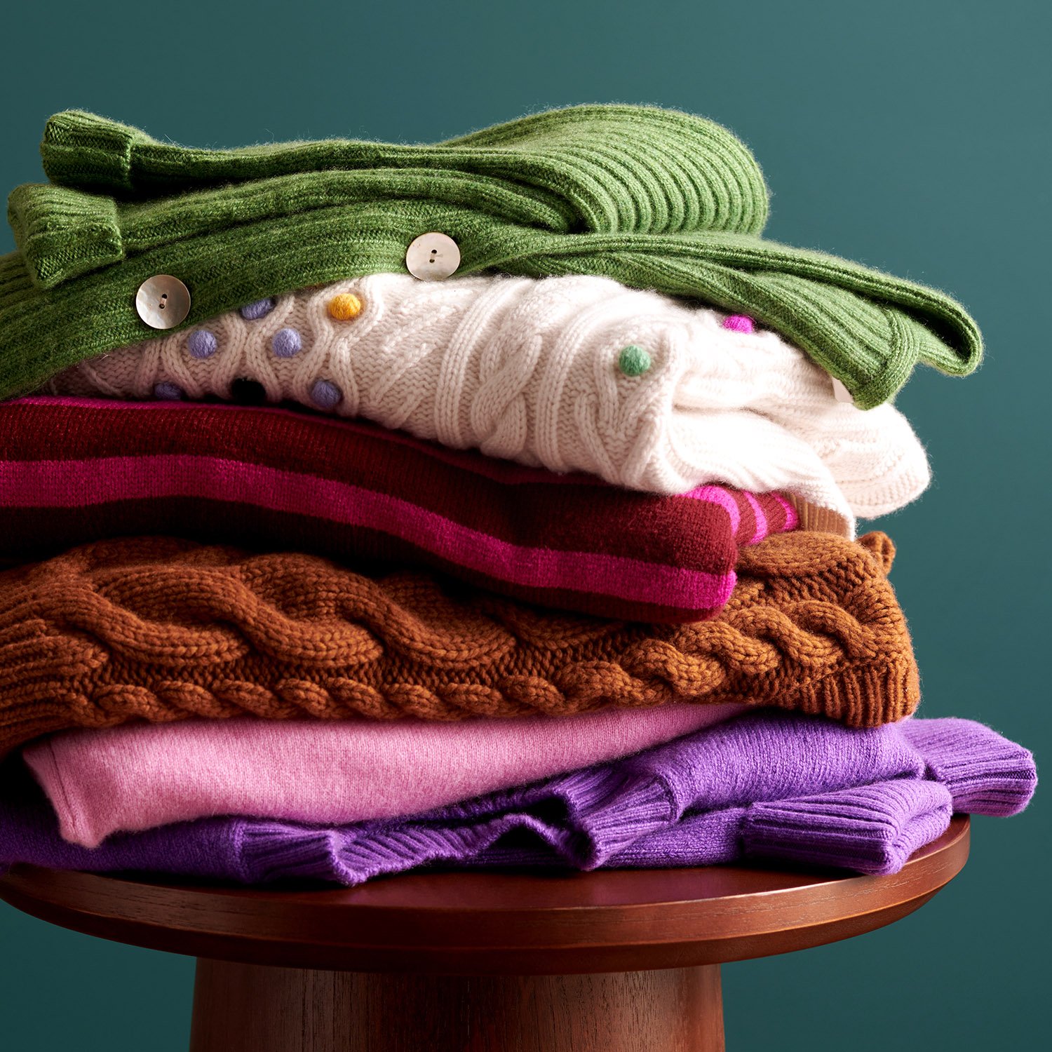 Stacked folded sweaters in green, white with polka dots, pink and magenta, orange, pink, purple, and lavender on a wooden table against a teal background.
