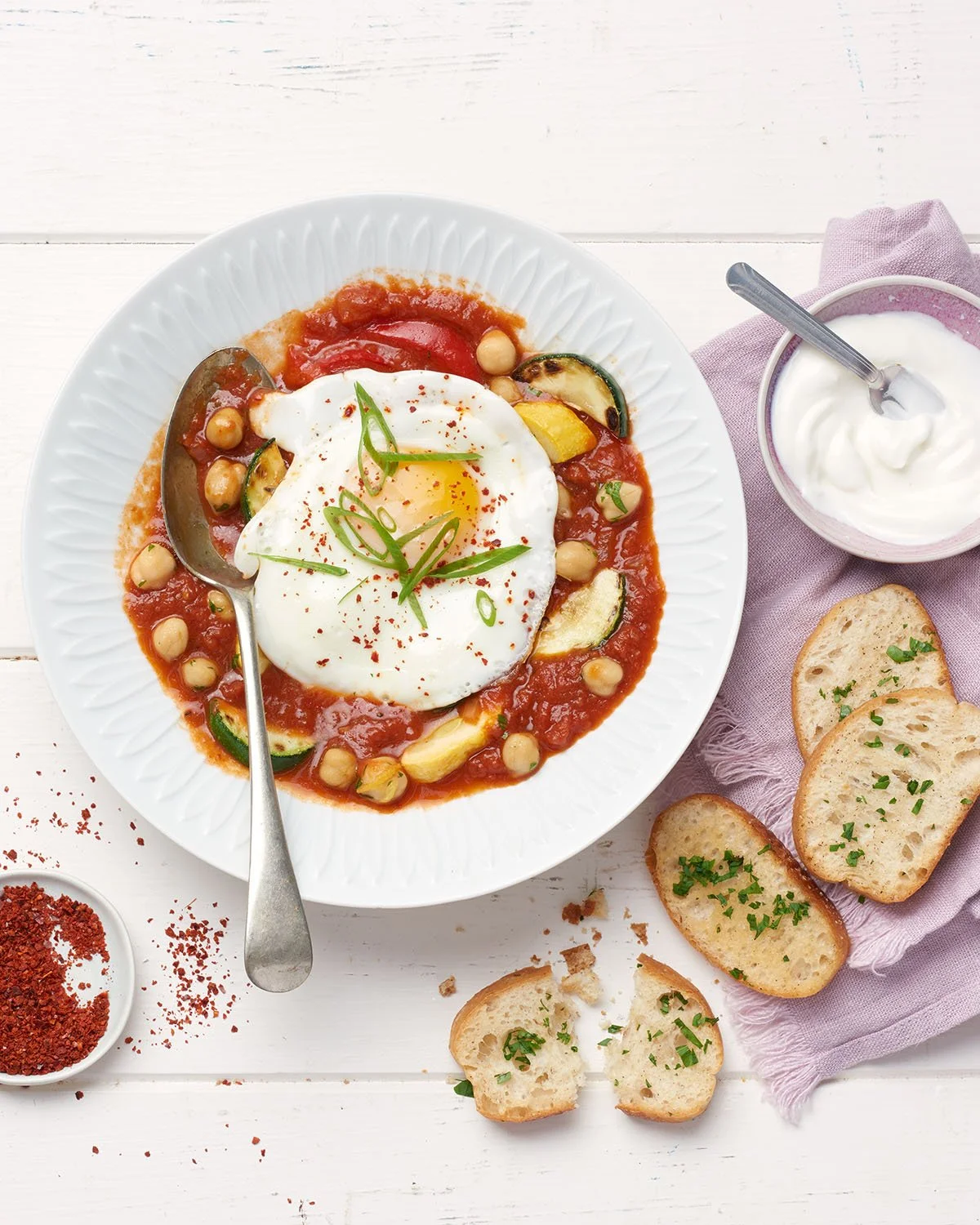 A plate of shakshuka with a poached egg, chickpeas, and vegetables in tomato sauce, garnished with green onions, with slices of bread and a bowl of yogurt on the side.