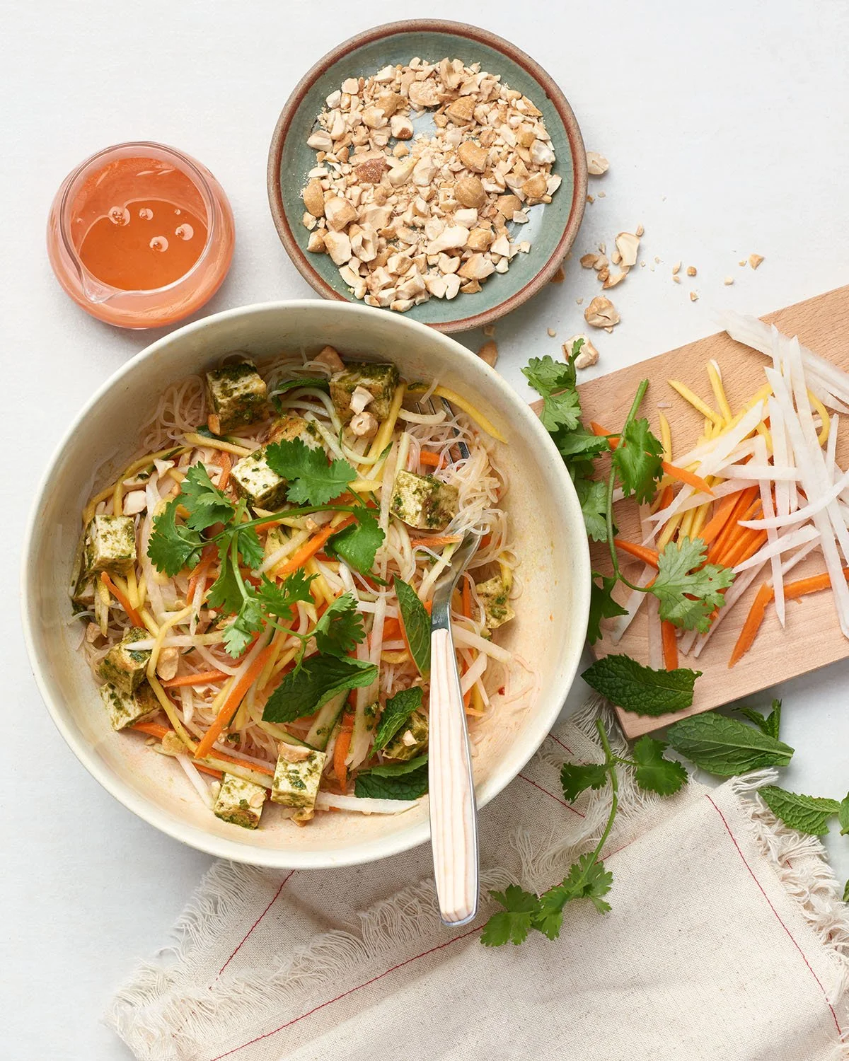 Bowl of Vietnamese-style rice noodle salad with tofu, shredded carrots, and herbs, with chopped peanuts and bean sprouts on a cutting board, and a glass of peach-colored juice on a white surface.