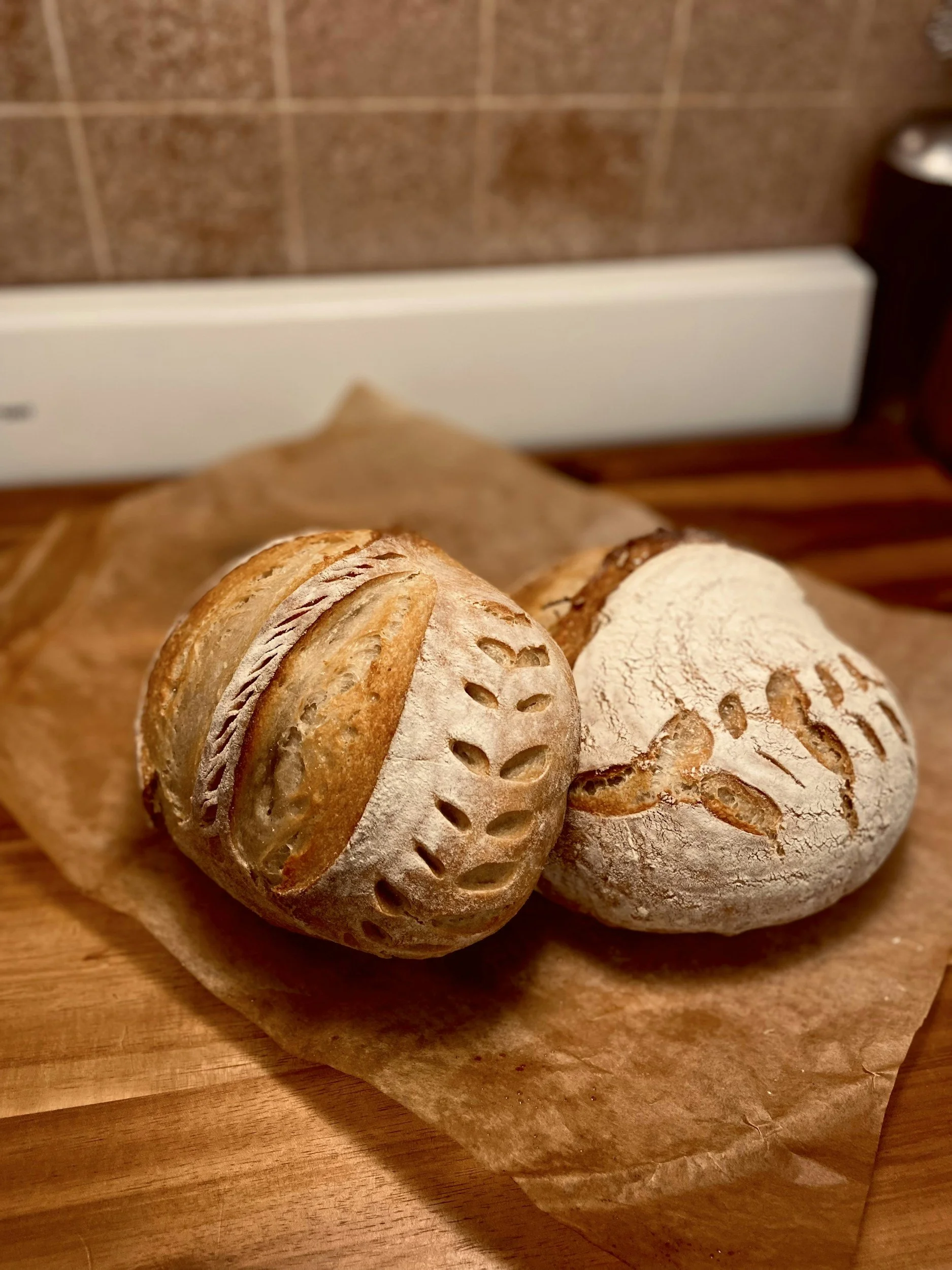 Two loaves of artisan bread on parchment paper on a wooden surface in a kitchen.
