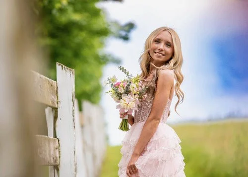 A young woman in a light pink dress holding a bouquet of flowers outdoors, smiling near a white fence with green trees and blue sky in the background.