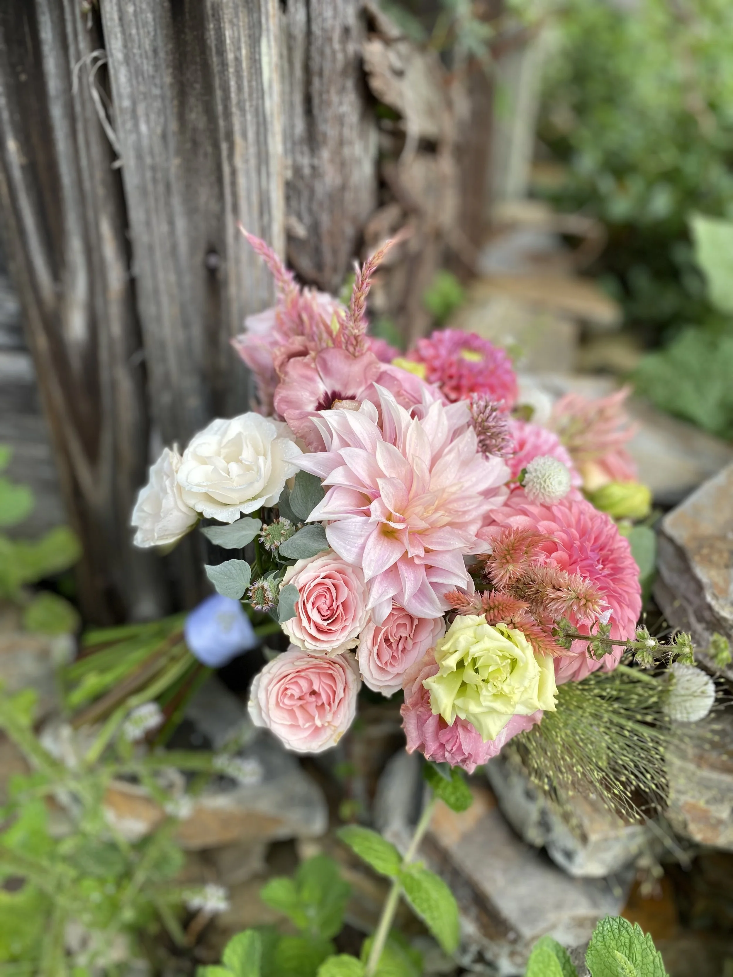 A bouquet of pink, white, and yellow flowers resting against a rustic wooden fence with green foliage in the background.