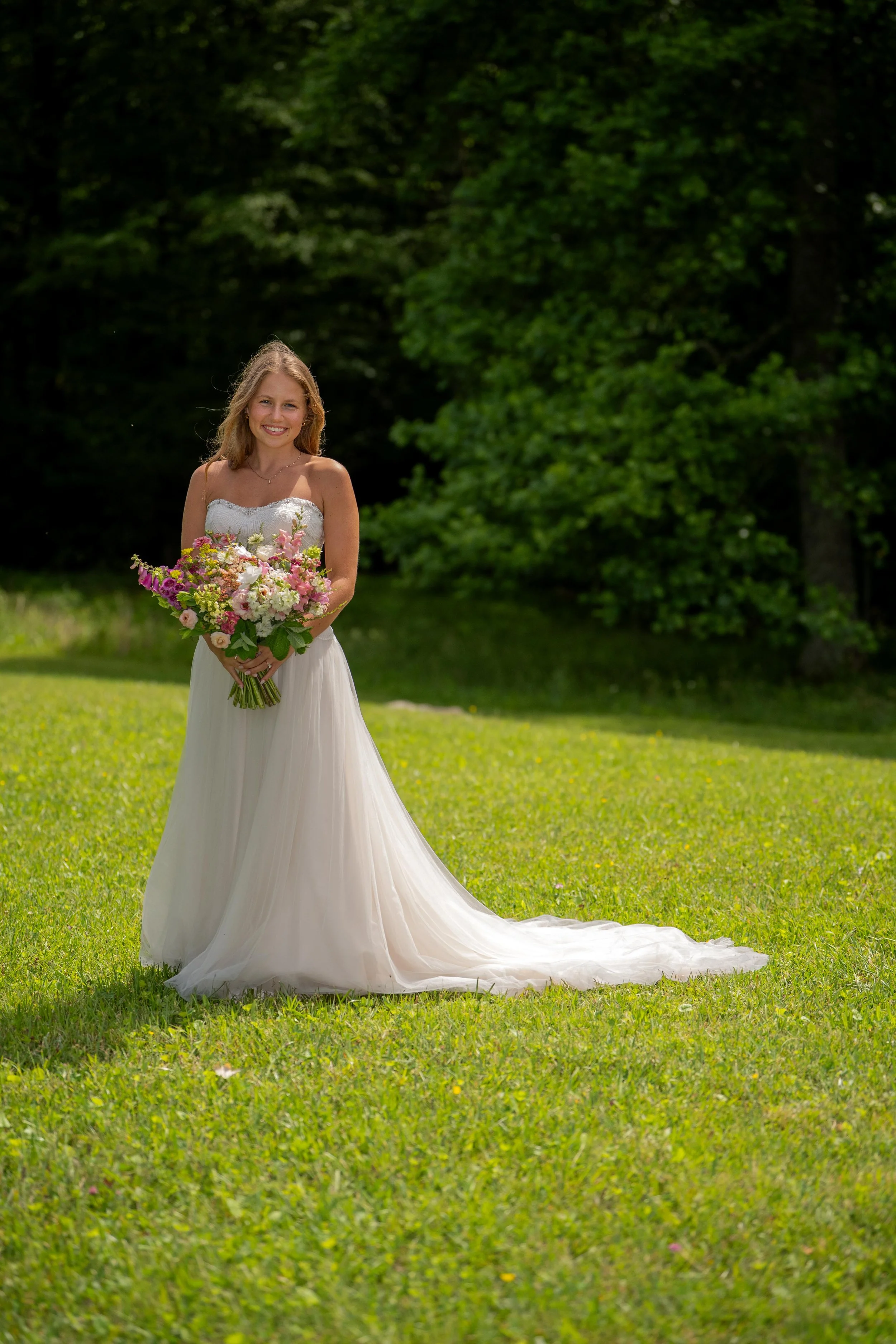 A smiling bride in a strapless wedding dress standing on a grassy field holding a colorful bouquet of flowers, with green trees in the background.