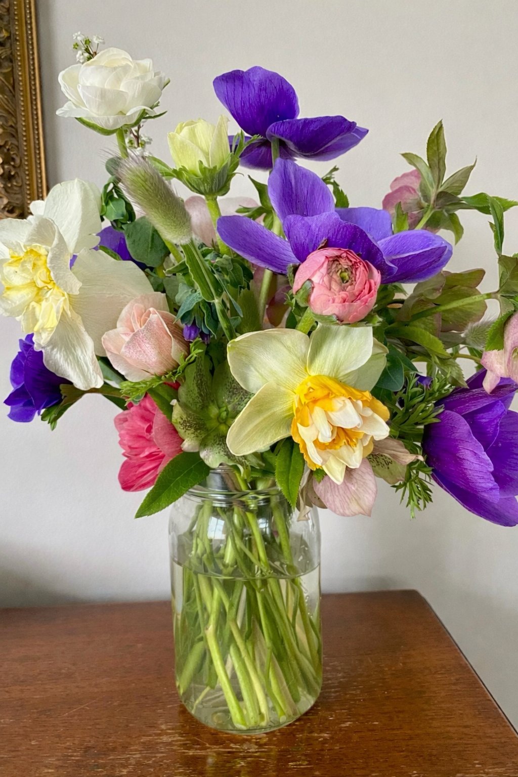 A colorful bouquet of mixed flowers including purple, pink, white, and yellow blooms in a clear glass vase on a wooden surface.