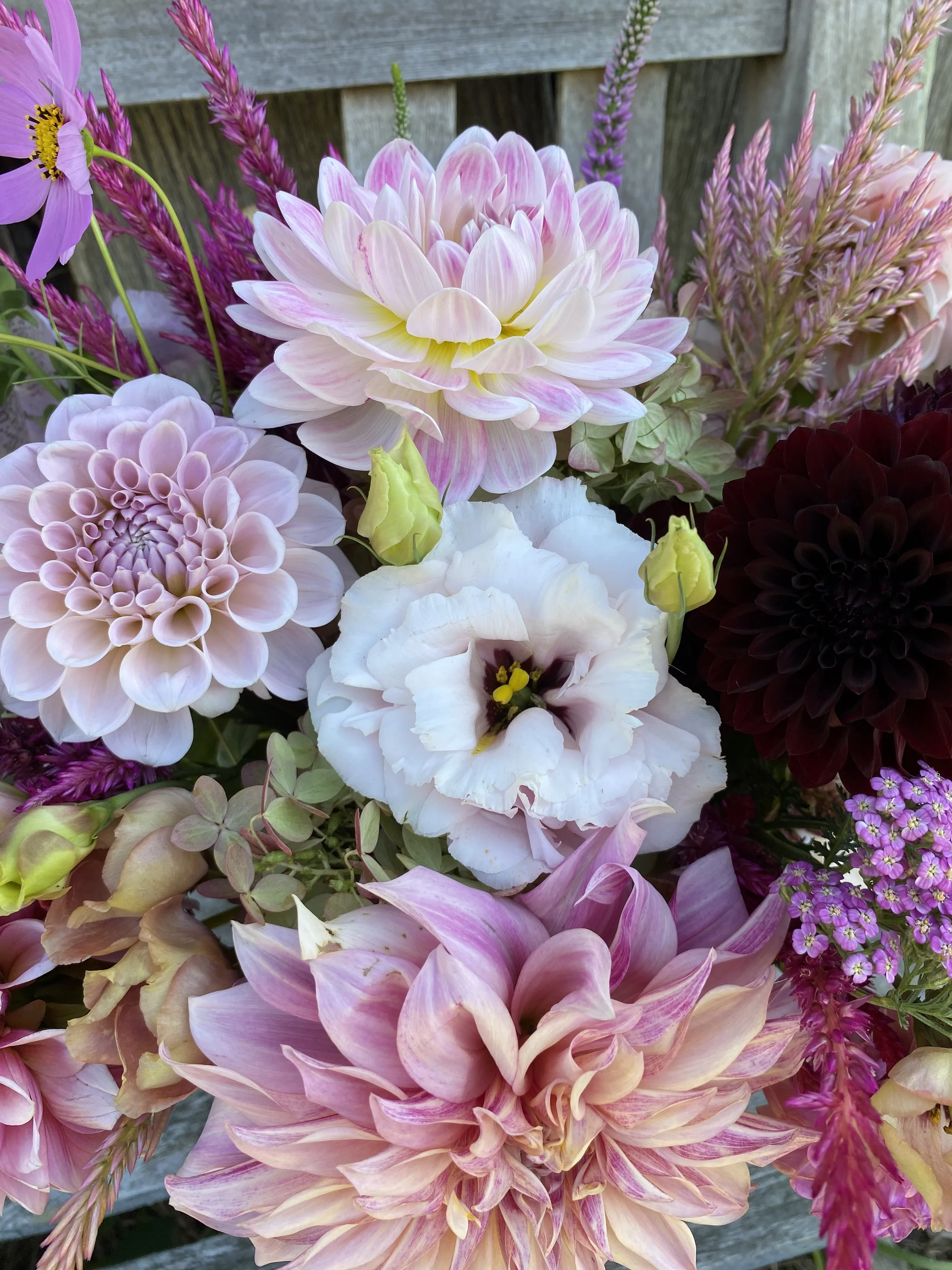 A colorful bouquet of various flowers including pink and white dahlias, a white lisianthus, purple and pink liatris, and small purple and pink flowers, placed against a wooden background.