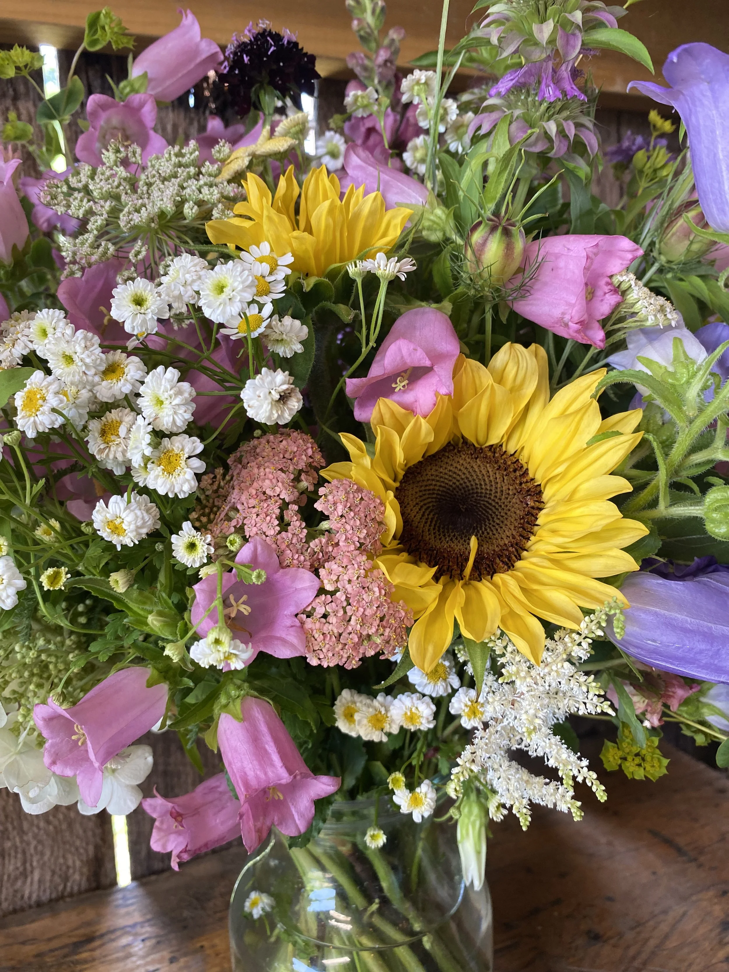 Colorful mixed flower bouquet in a glass vase, including sunflowers, pink and purple flowers, and small white flowers, on a wooden table.