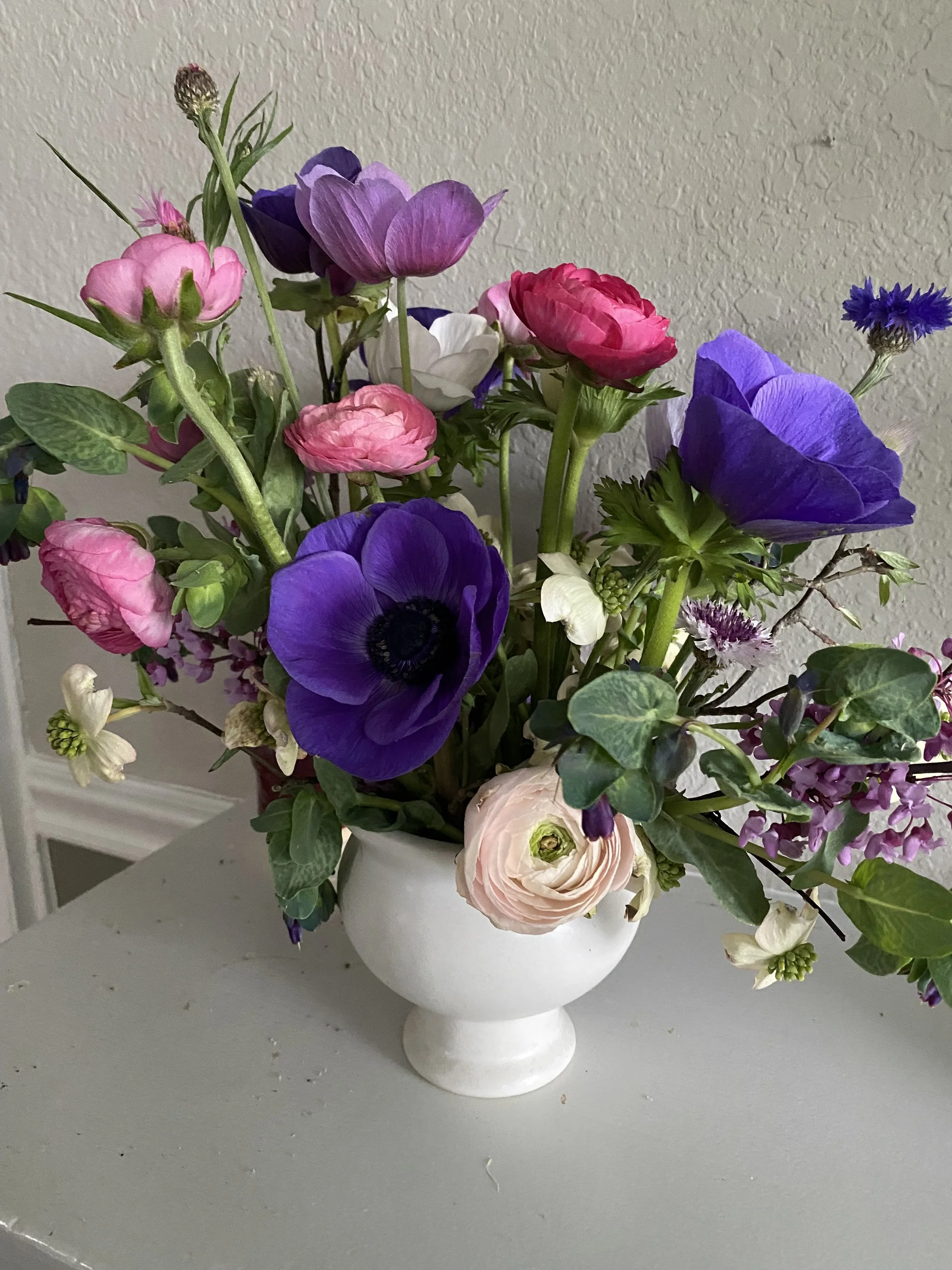 A white vase with a bouquet of colorful flowers including purple anemones, pink ranunculus, and other mixed blooms placed on a white surface against a textured beige wall.