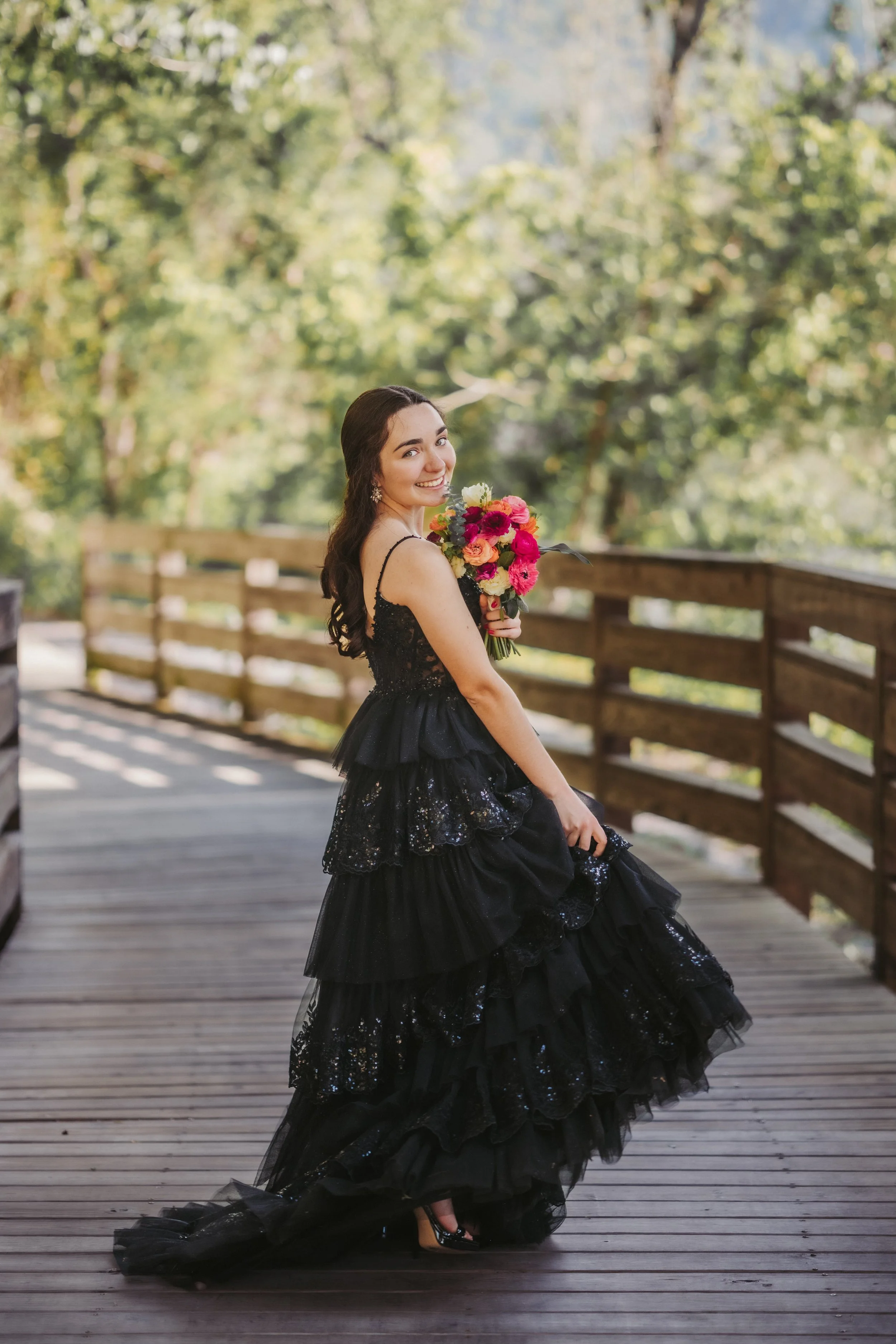 A woman in a black, sequined, ruffled gown holding a colorful bouquet on a wooden bridge surrounded by green trees.