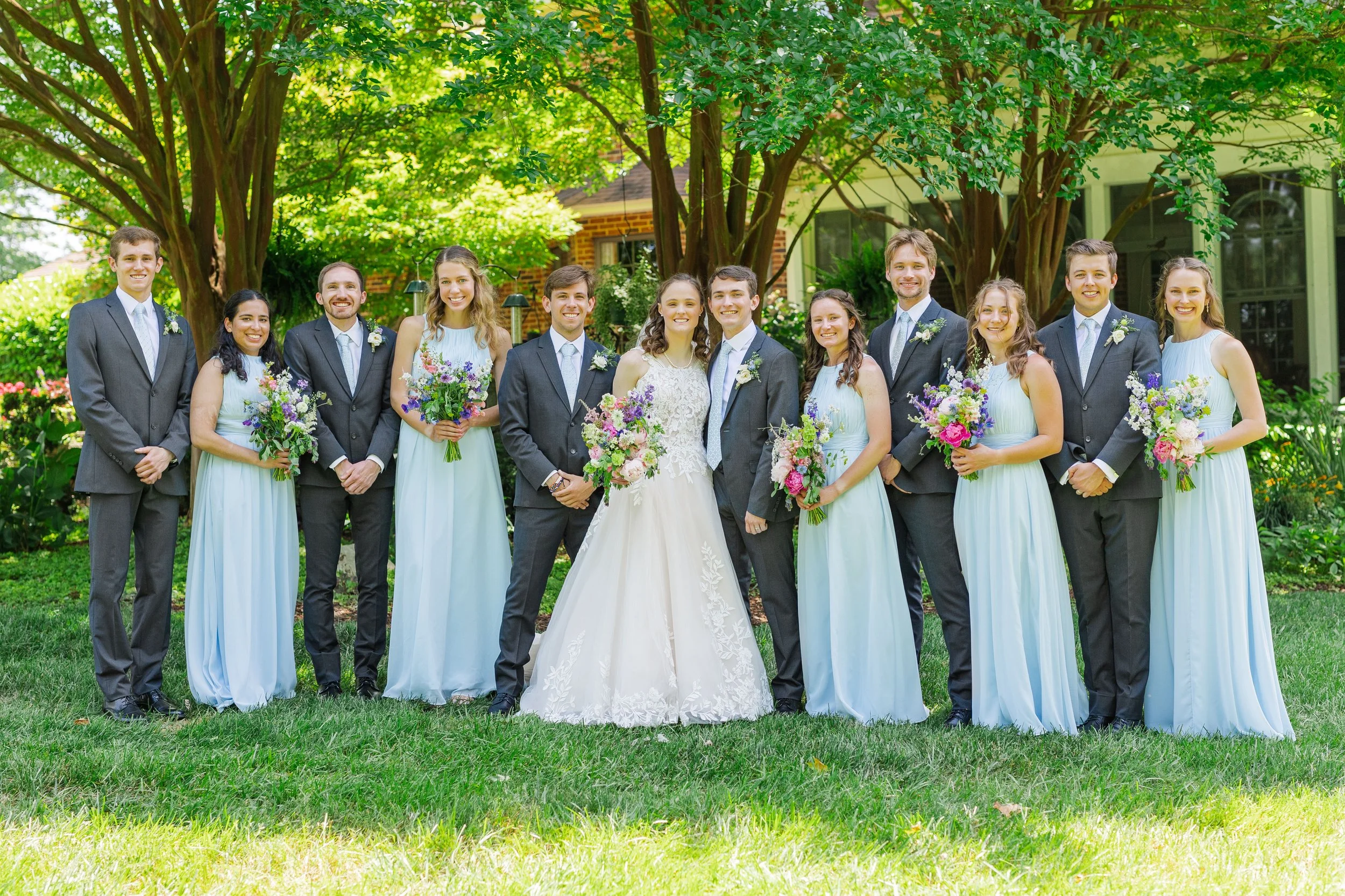 A bride and groom with their wedding party standing outdoors on grass with trees and a house in the background. The wedding party includes six women in light blue dresses holding bouquets and six men in gray suits holding bouquets.
