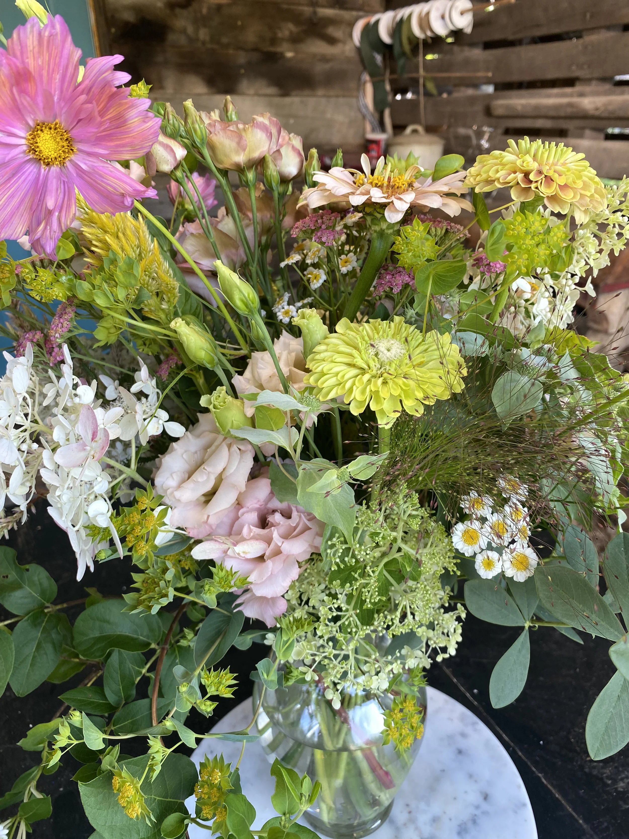 A mixed bouquet of various colorful flowers including pink, white, yellow, and cream flowers in a glass vase on a white marble surface, with a rustic wooden background.