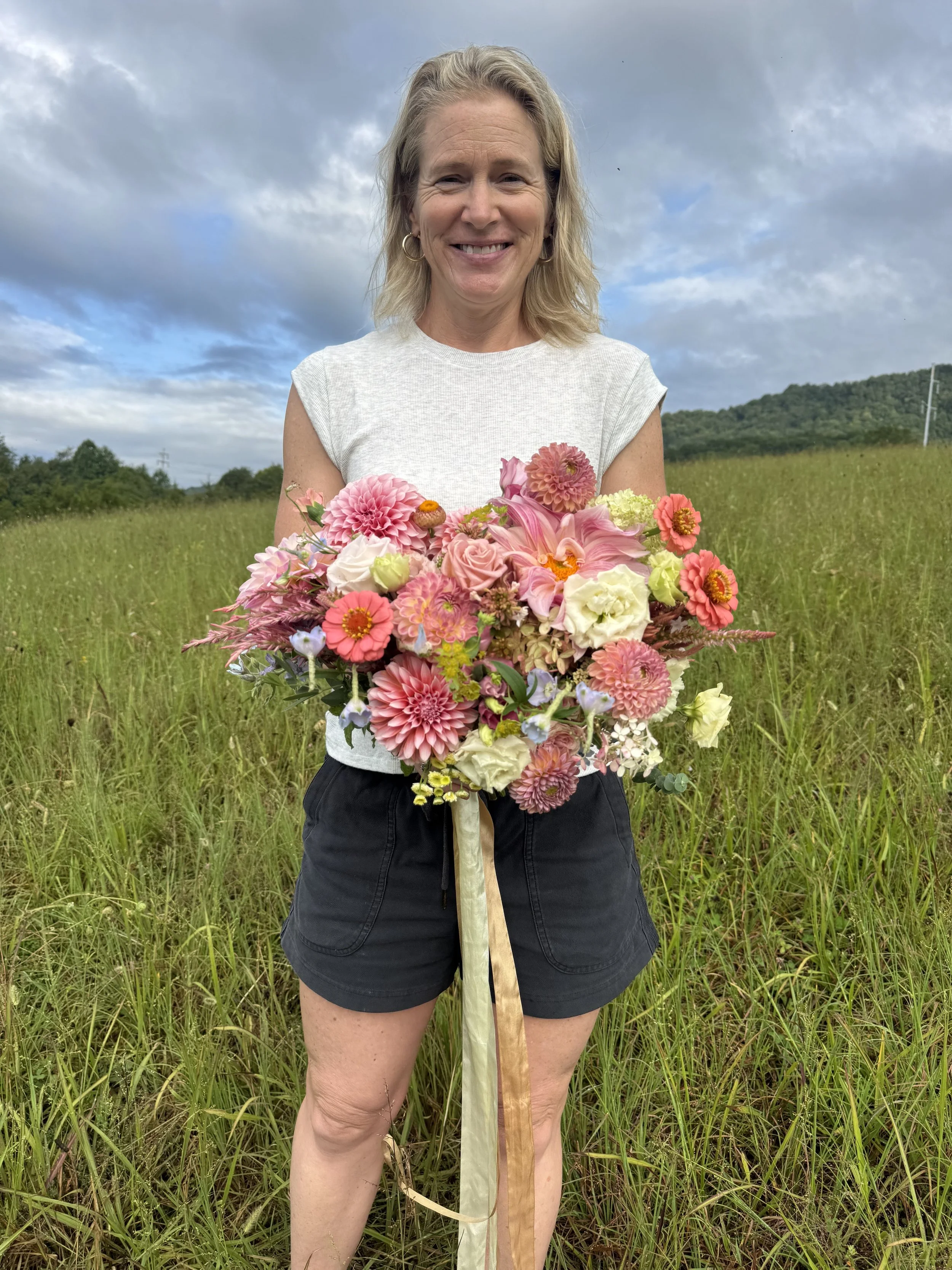 Woman holding a bouquet of pink and white flowers standing in a grassy field with a cloudy sky and distant hills in the background.