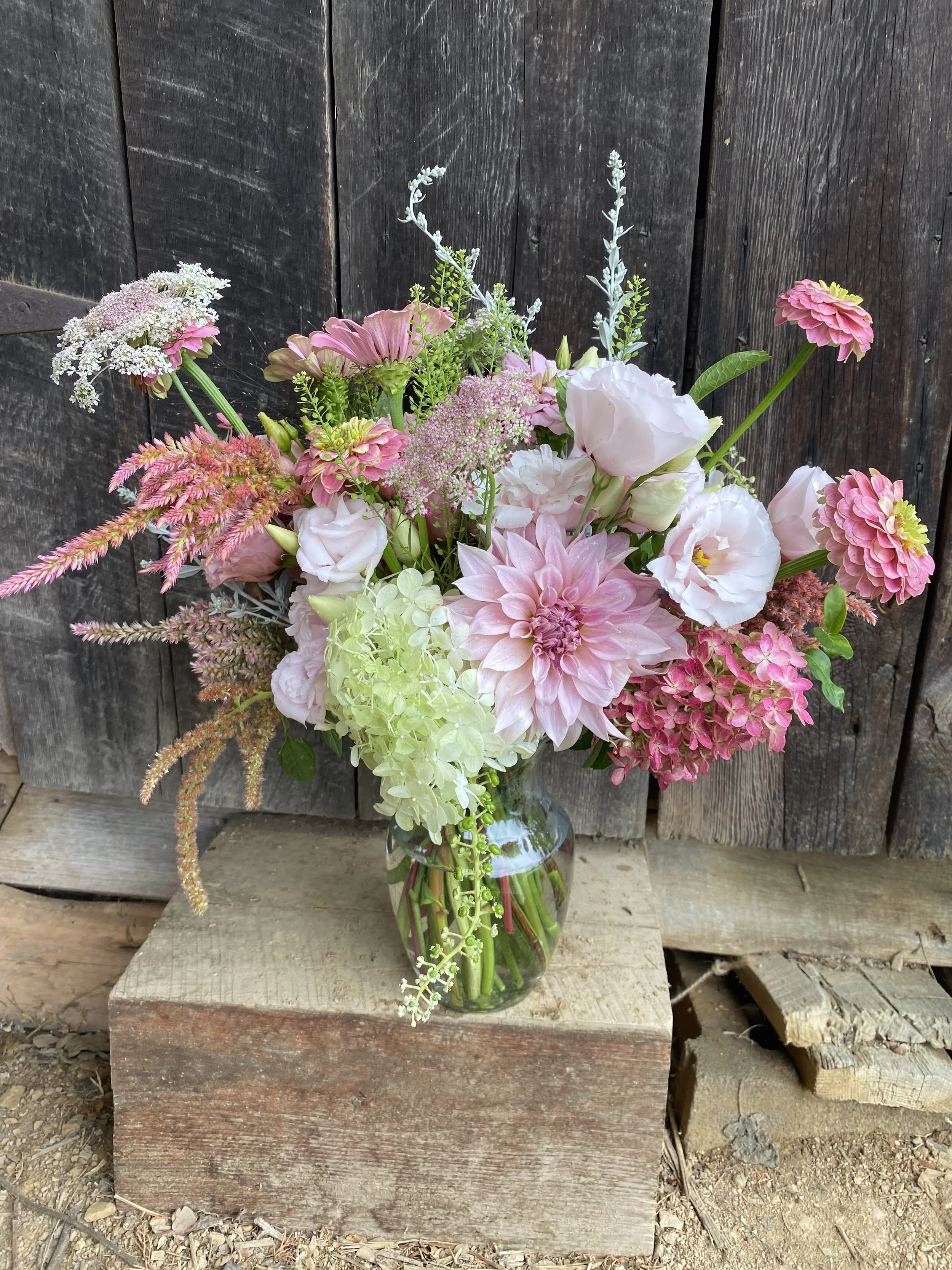 A colorful bouquet of pink and white flowers in a clear glass vase, placed on a wooden pedestal against a rustic dark wooden backdrop.