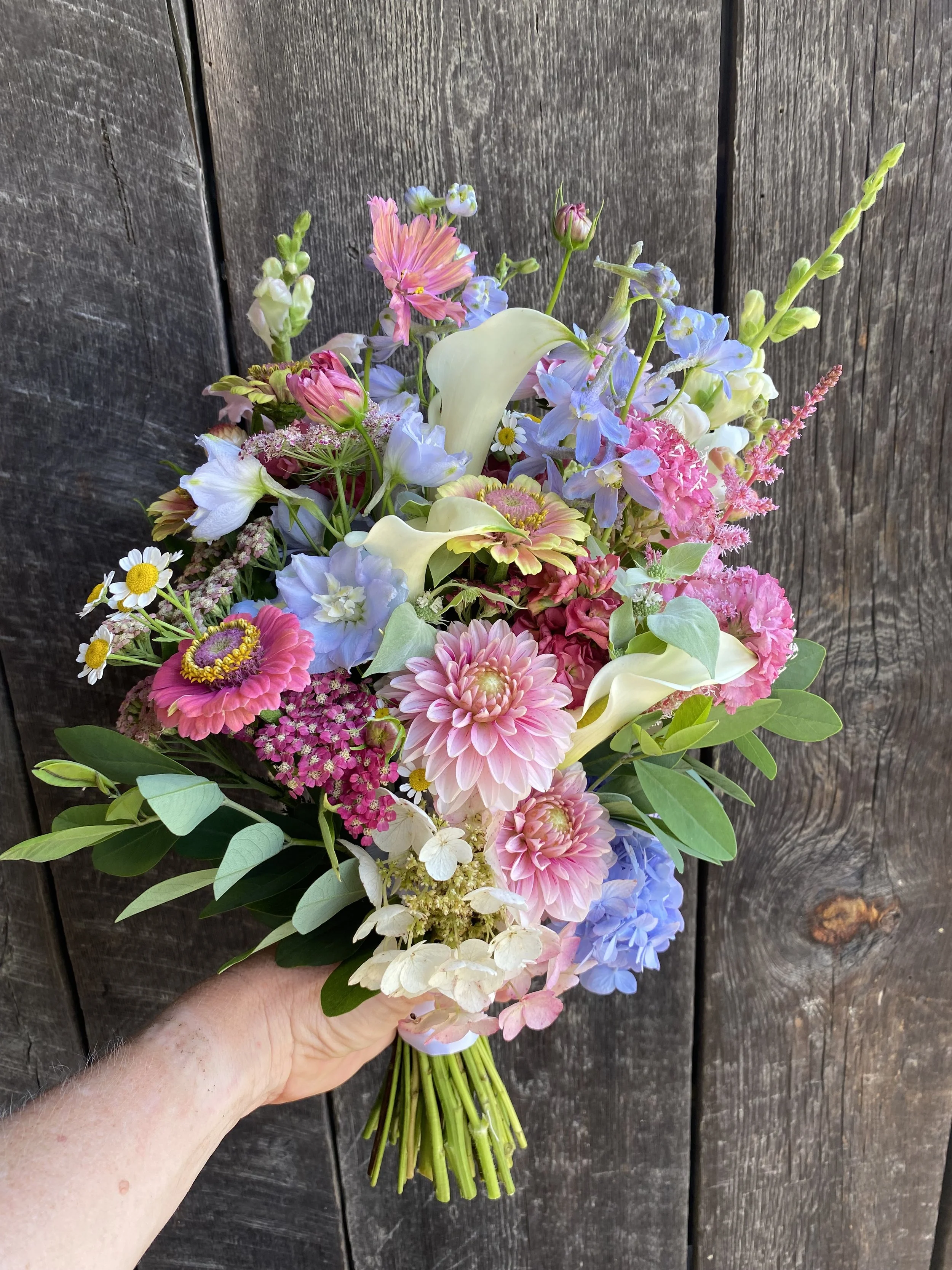 A hand holding a colorful bouquet of mixed flowers including dahlias, calla lilies, daisies, and other blooms, against a background of dark wooden panels.
