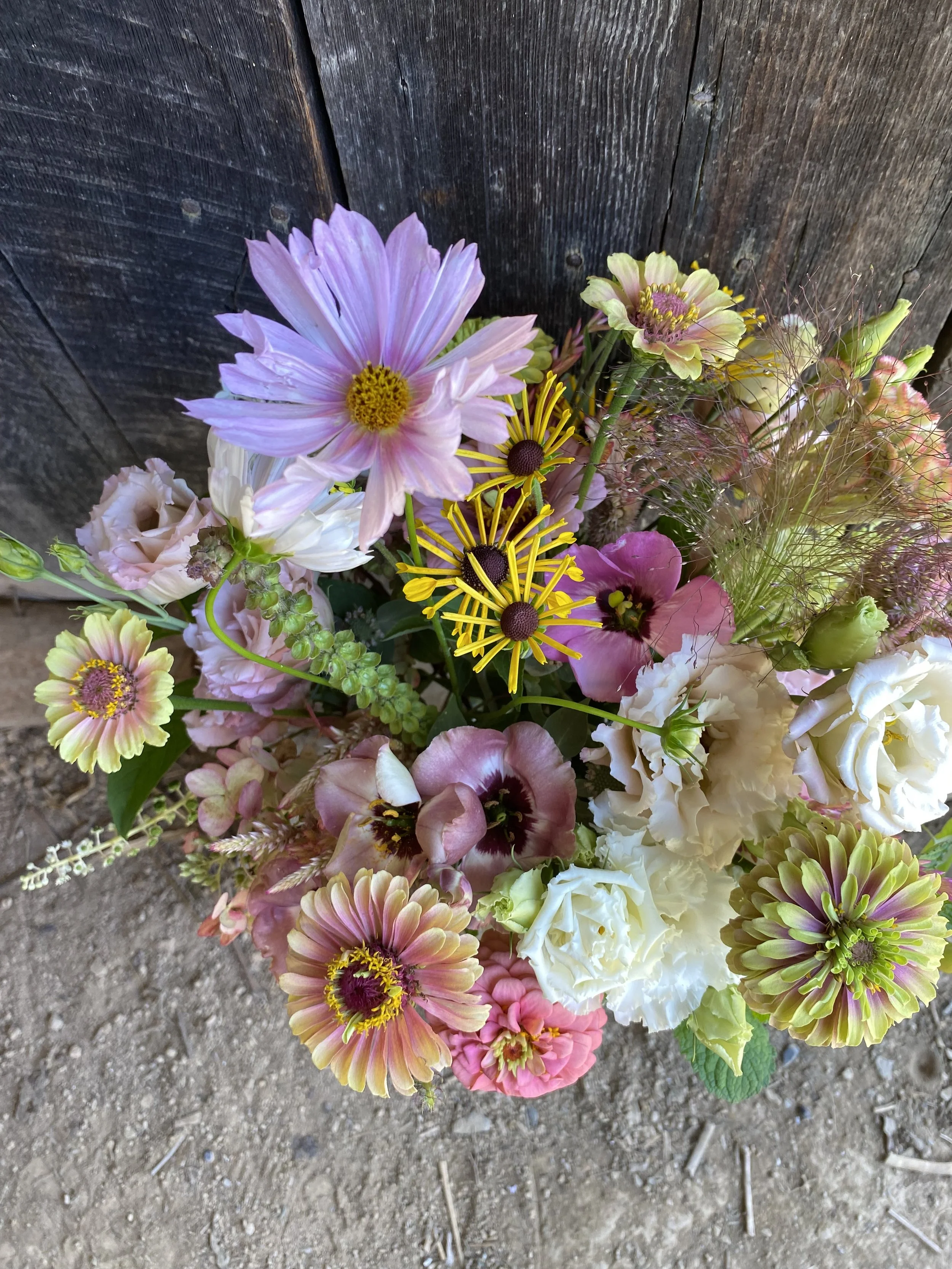 Colorful bouquet of various fresh flowers, including pink and white blooms, with a wooden background and dirt ground.