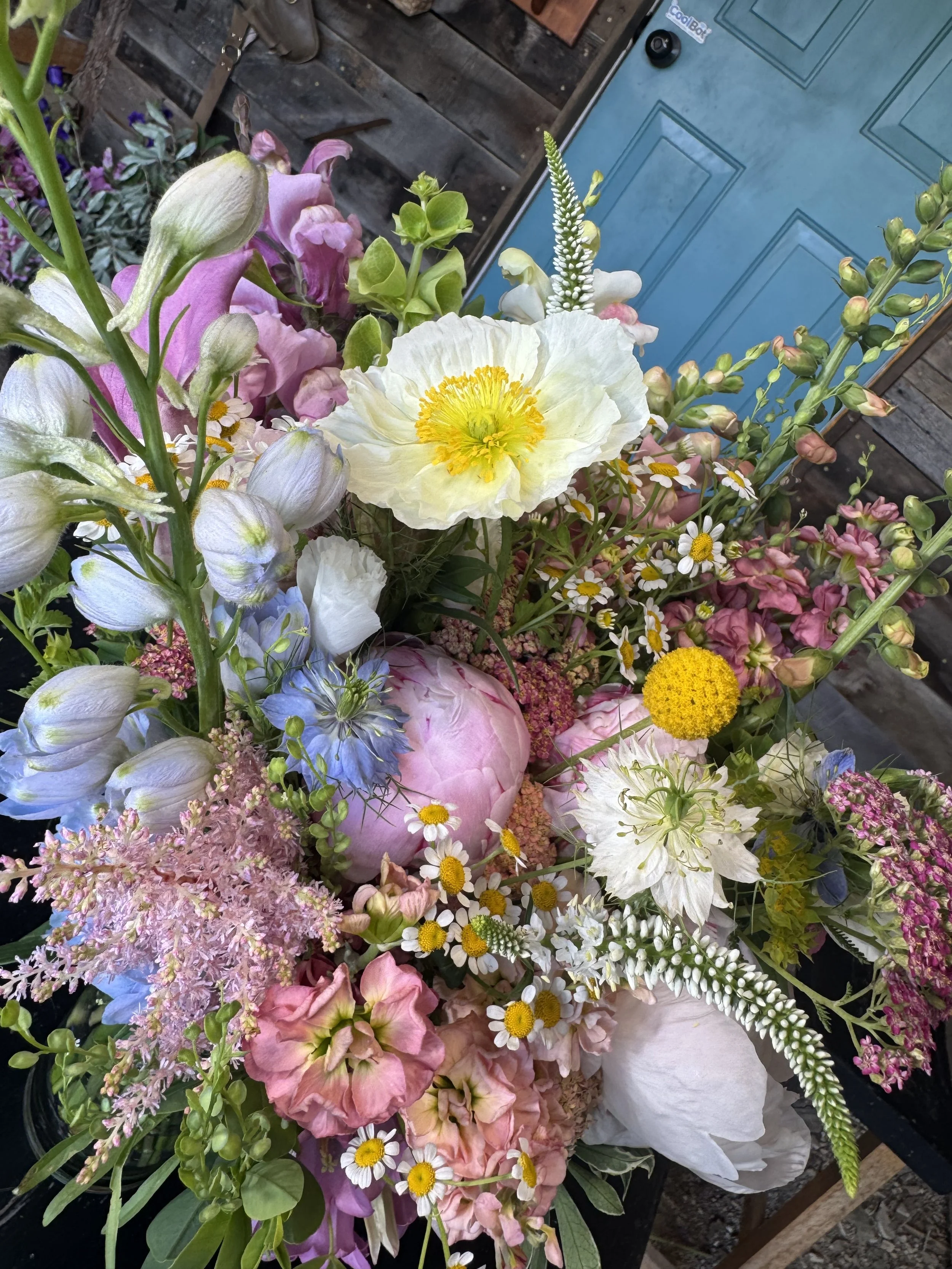 A colorful arrangement of various flowers, including pink, white, blue, and yellow blossoms, in front of a rustic wooden background and a blue door.