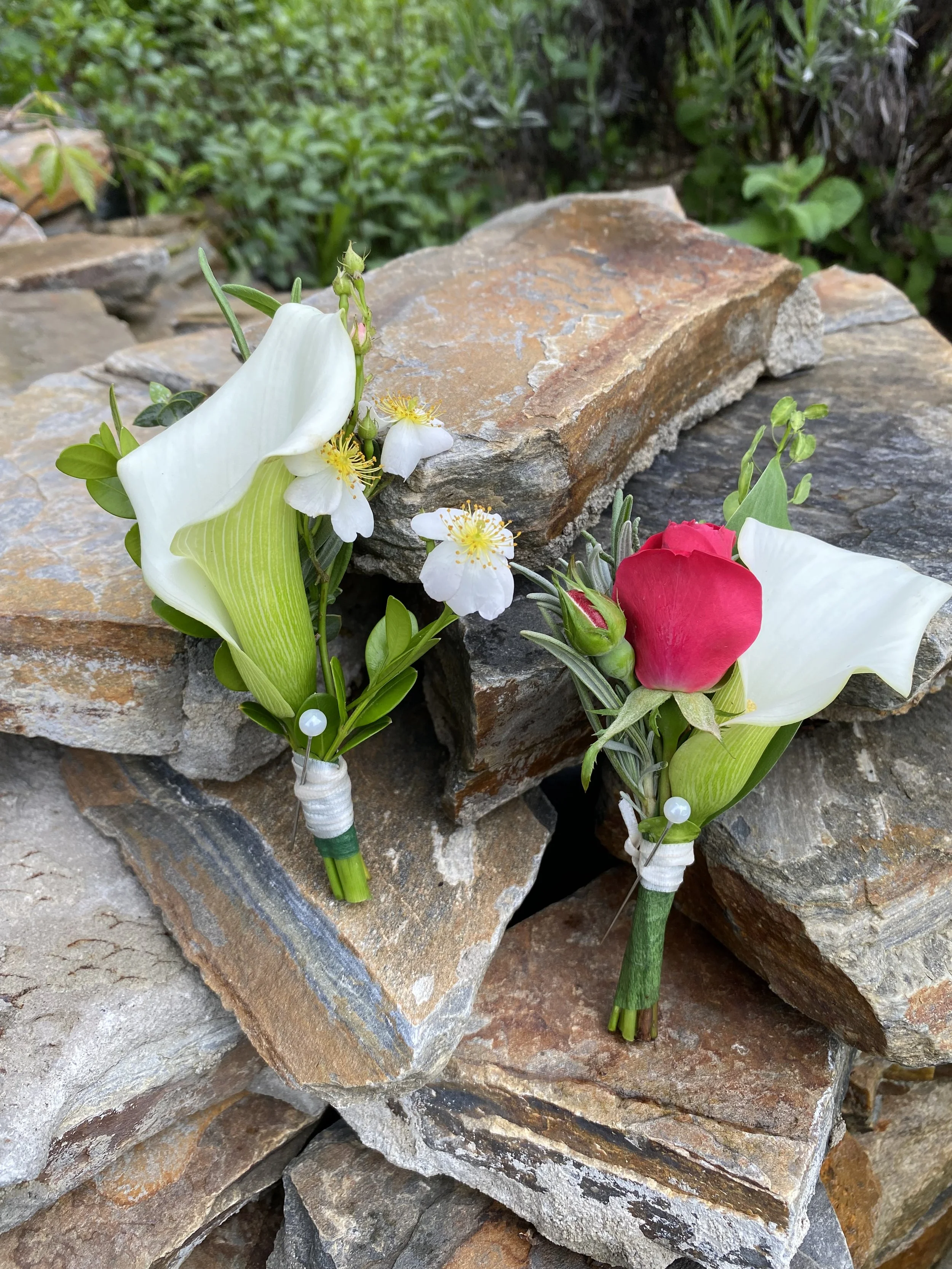 Two small floral arrangements with white lilies and small white flowers, one with a red rosebud, placed on stacked stones.