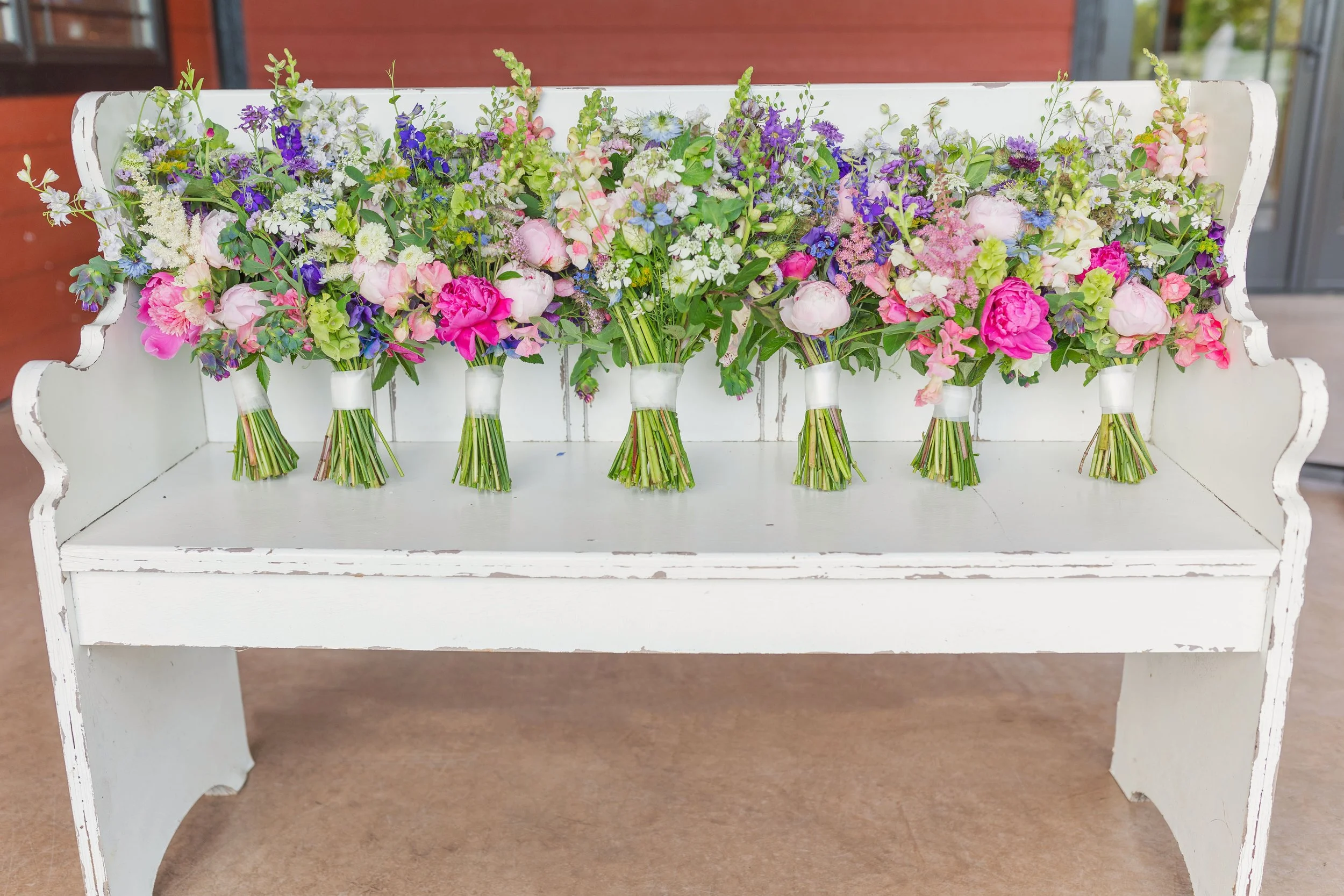 Seven bouquets of mixed pink, purple, white, and green flowers displayed on a white wooden bench.