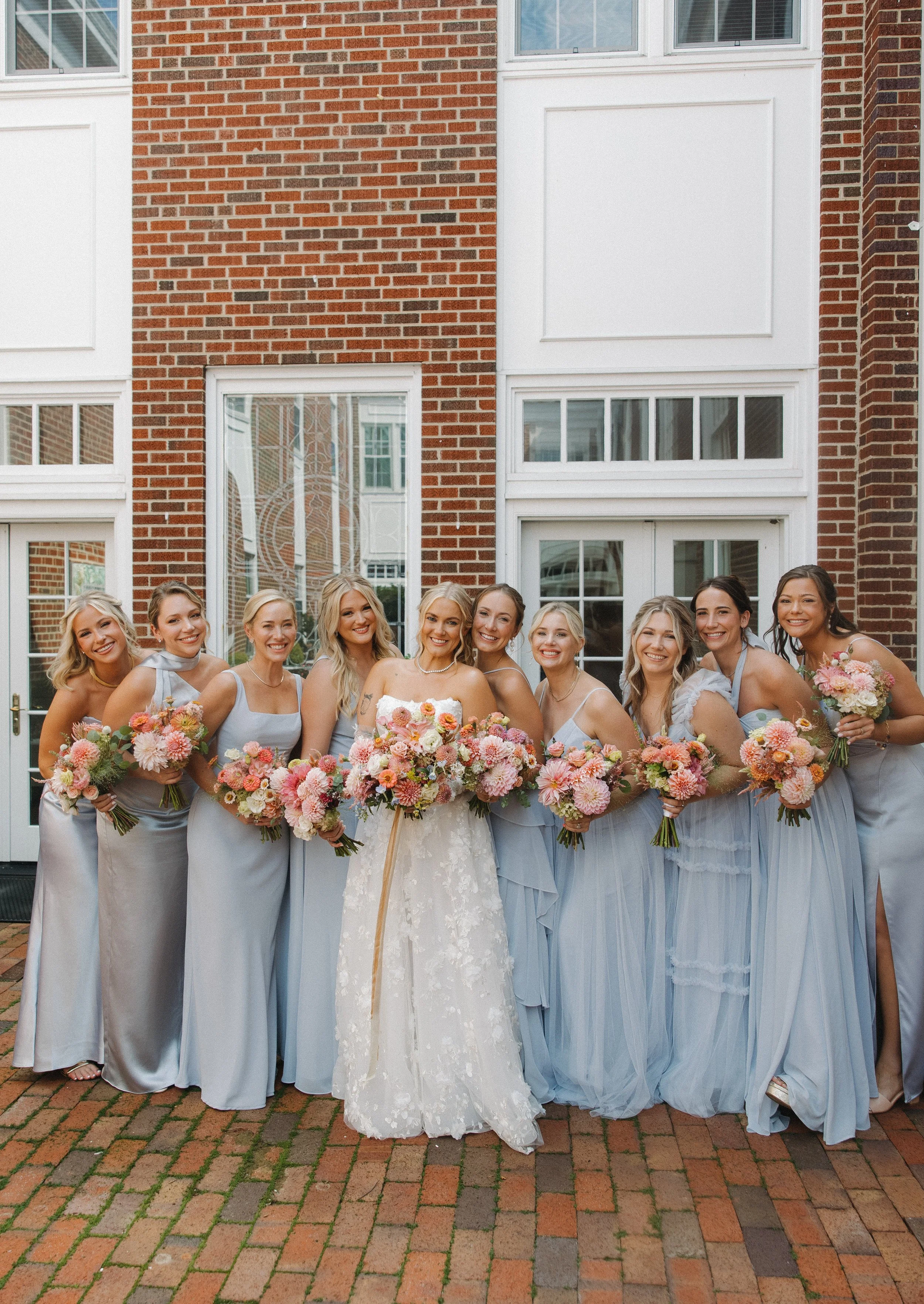 A group of eleven women in pastel dresses holding bouquets of pink and peach flowers, standing in front of a brick building with large white-framed windows, smiling for a group photo at a wedding celebration.