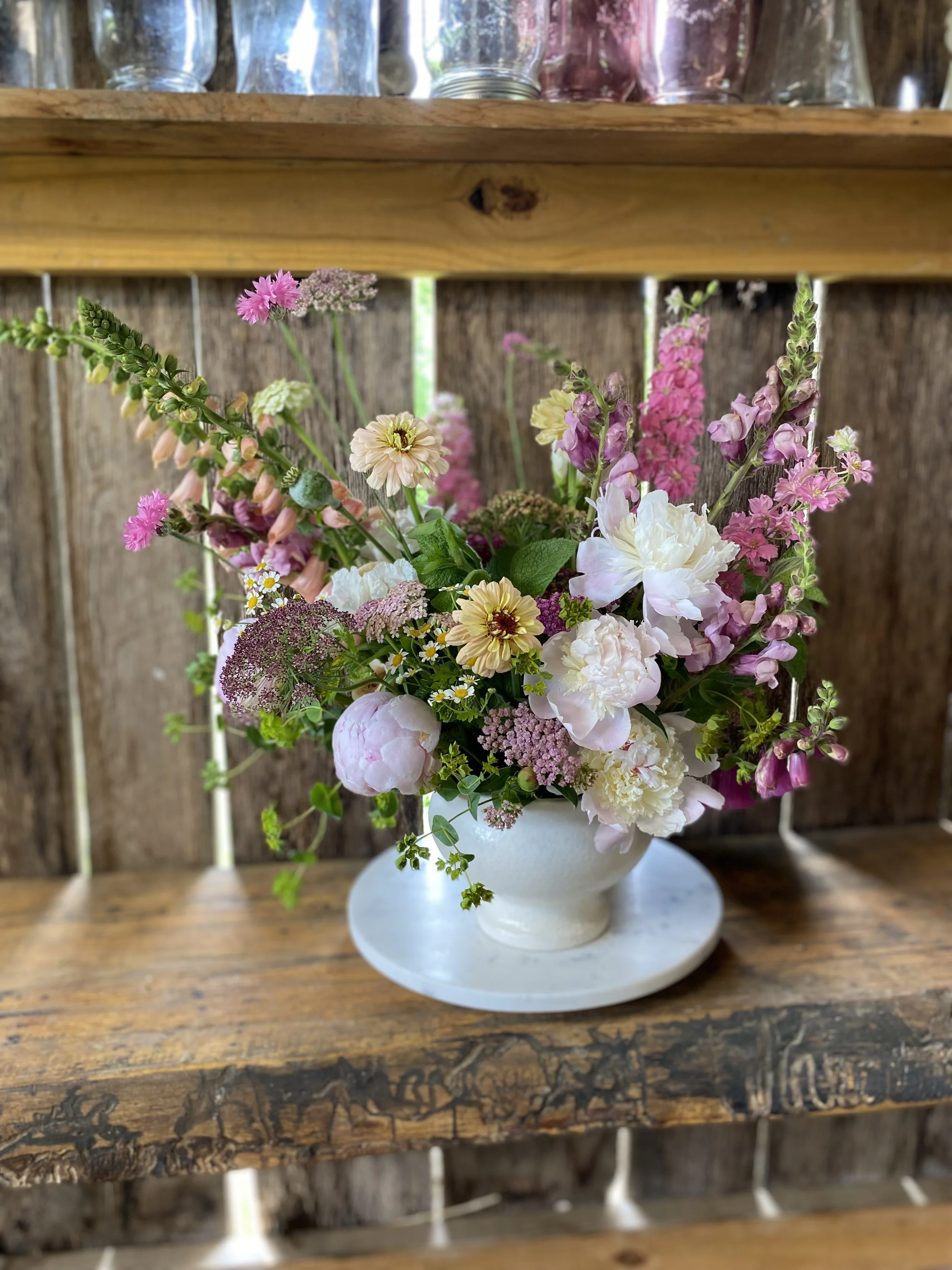 A white flower vase with a mix of pink, white, and yellow flowers on a wooden shelf.