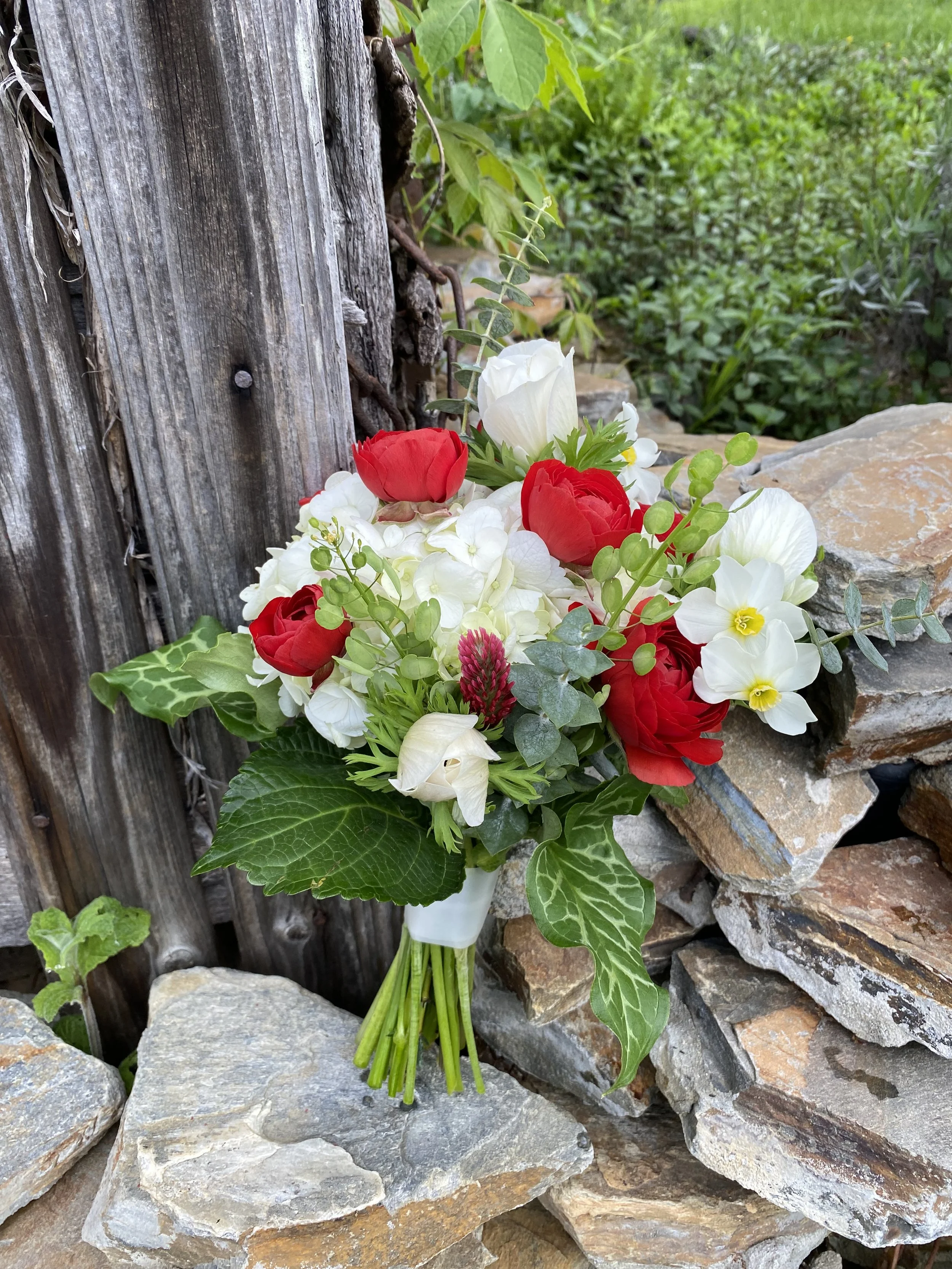 A bouquet of white, red, and cream flowers with green leaves, resting against a rustic stone and wood background.