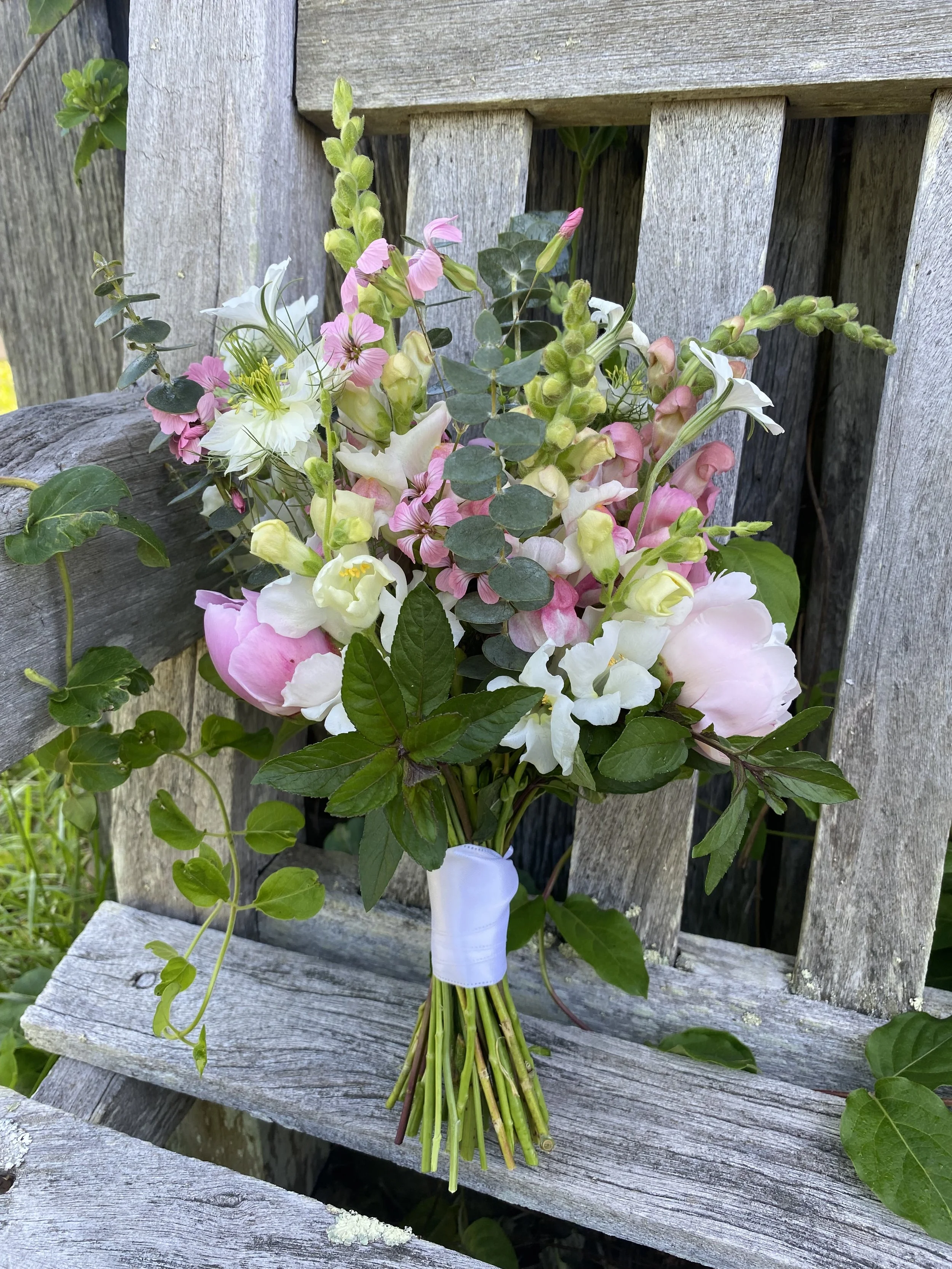 A bouquet of pink, white, and green flowers with green leaves, tied with a white ribbon, placed on a weathered wooden bench outdoors.
