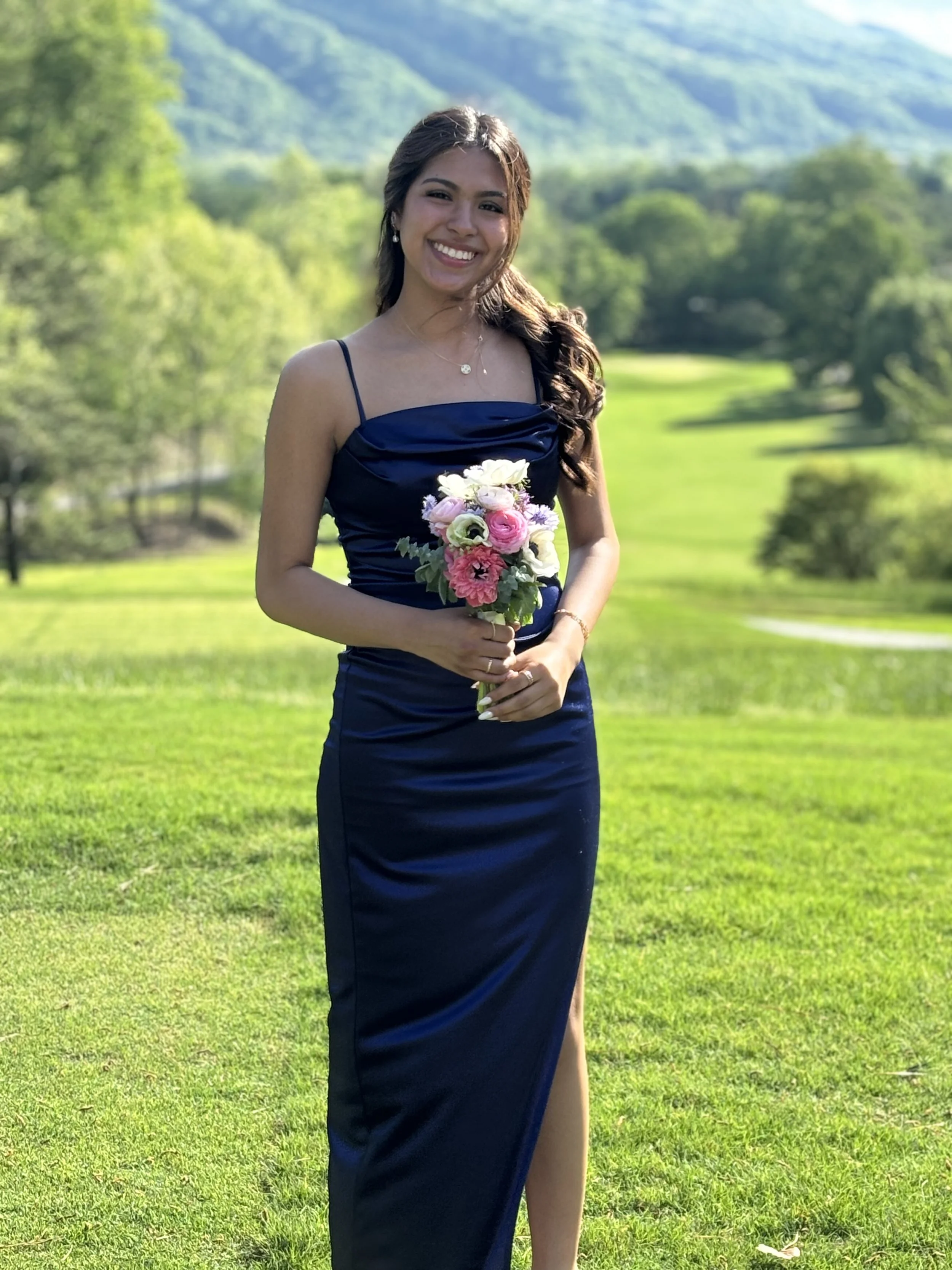 A young woman in a navy blue dress holding a bouquet of pink and white flowers standing on a grassy field with trees and mountains in the background.