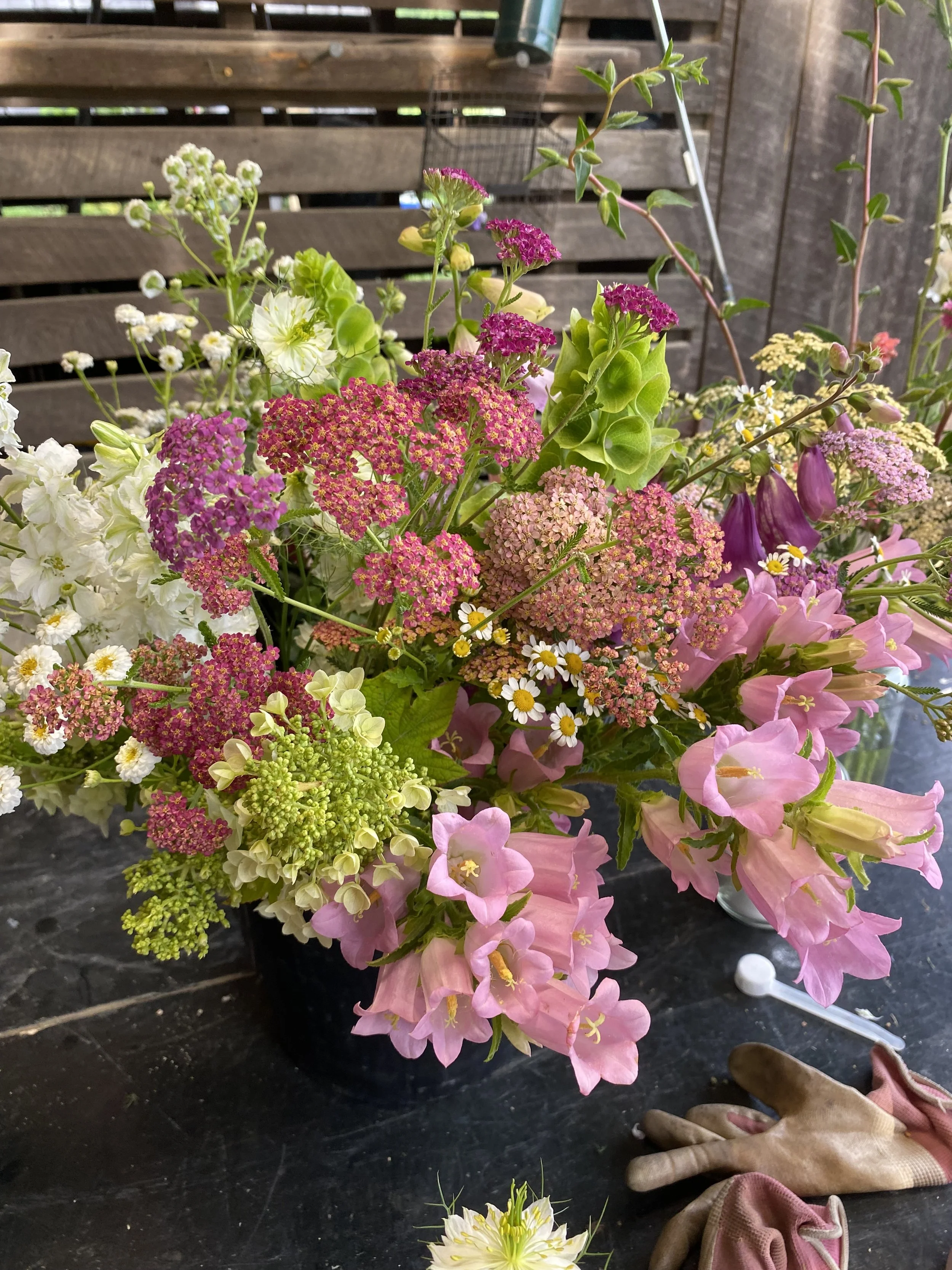 A vibrant bouquet of various colorful flowers including pink, white, purple, and green blooms, arranged in a black container on a black surface, with gardening gloves and a cotton swab nearby.