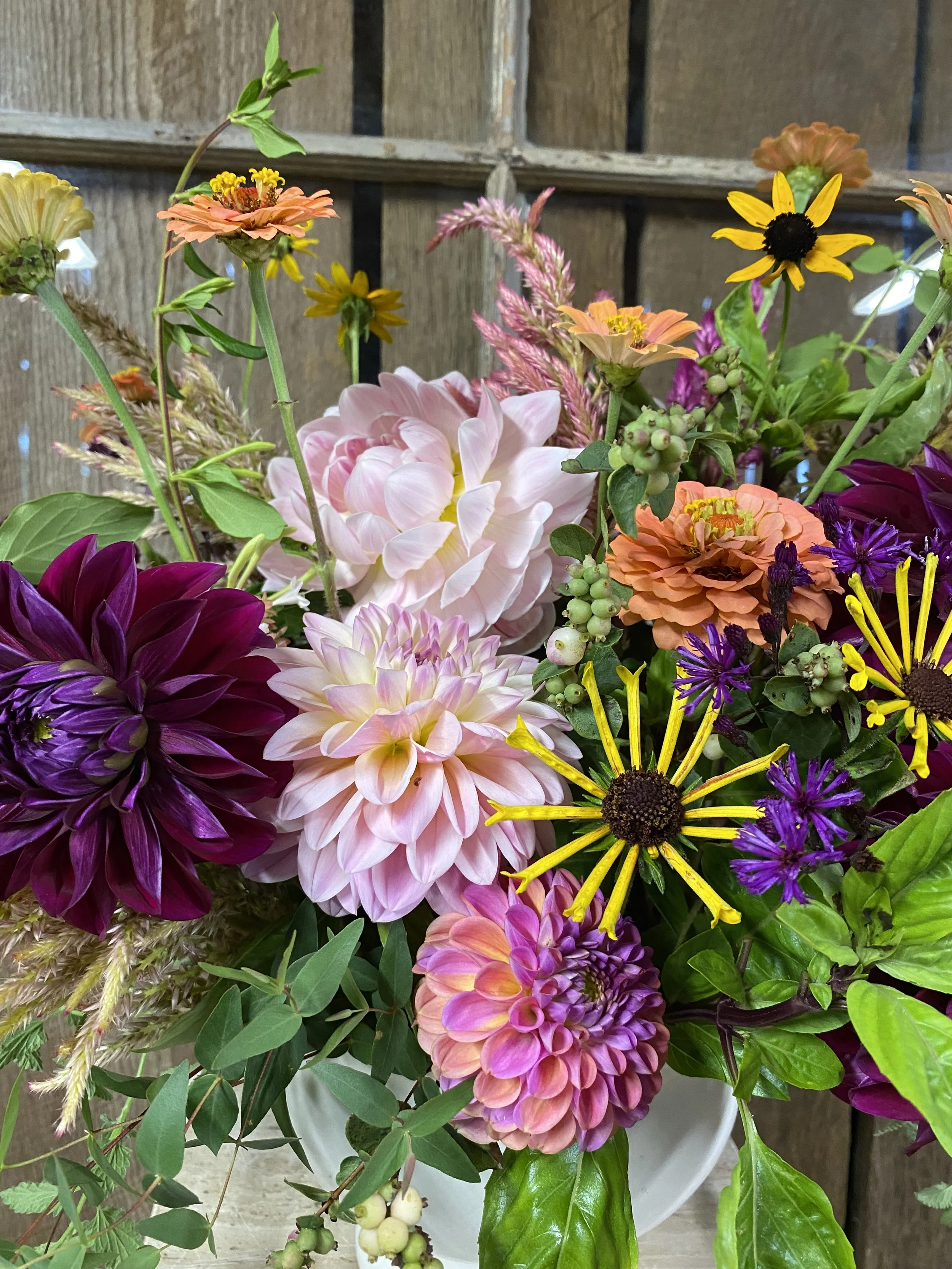 Colorful mixed flower bouquet with dahlias, black-eyed Susans, purple and yellow asters, and other vibrant flowers in a white vase against a wooden background.
