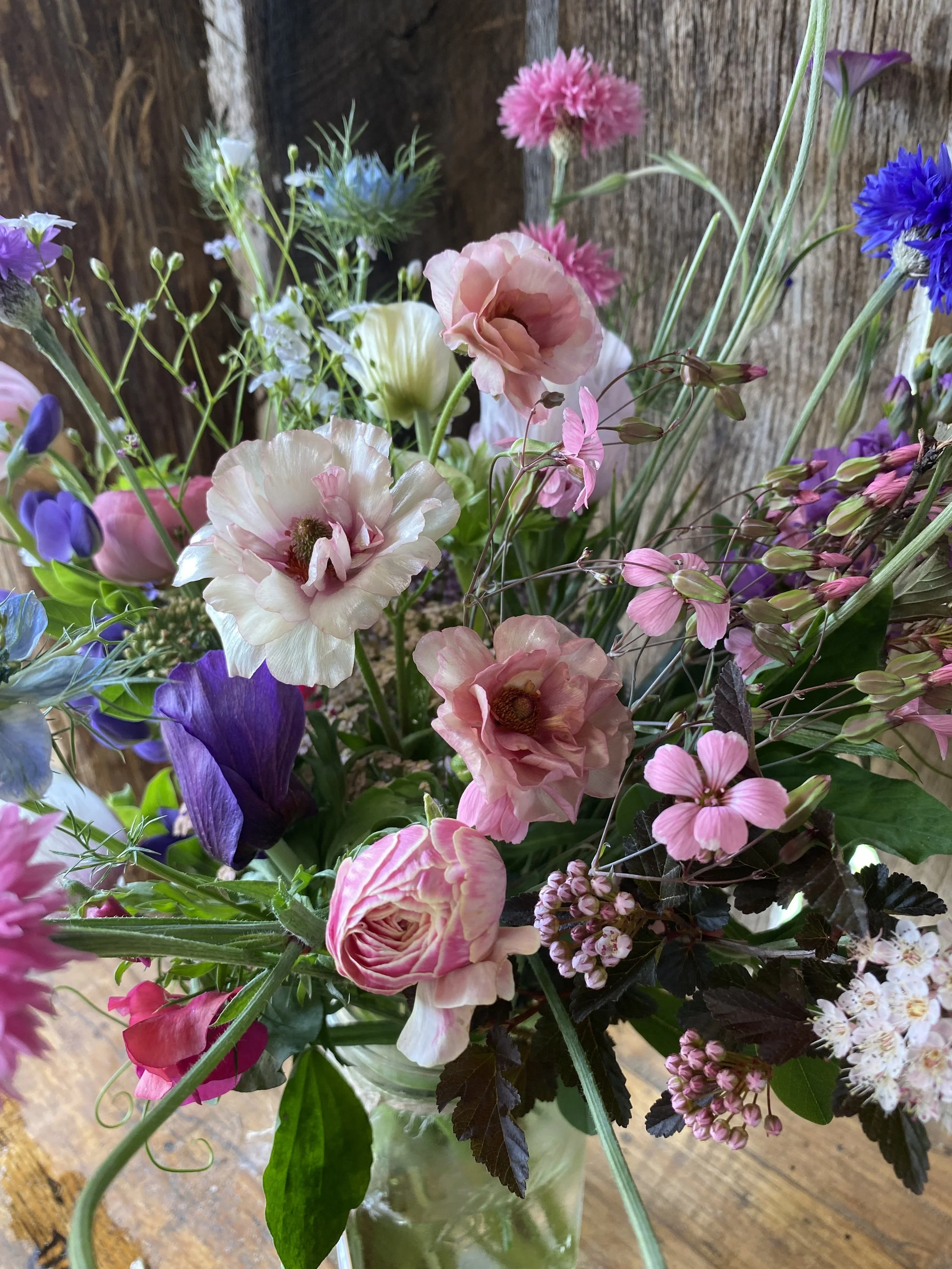 Colorful bouquet of mixed flowers including pink and purple blooms in a glass vase on a wooden surface, with a wooden background.