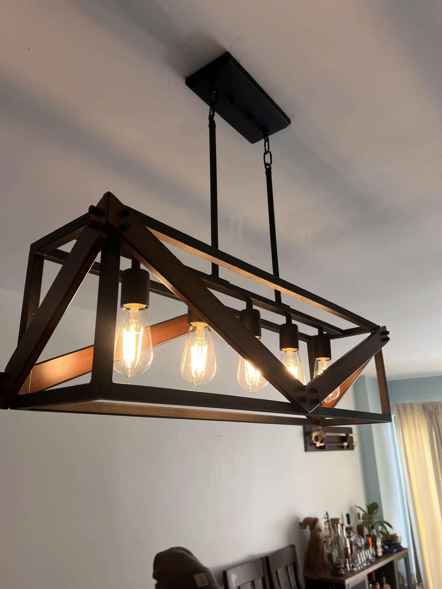 Modern rectangular black and wood chandelier with five exposed light bulbs hanging from the ceiling in a room with light-colored walls and a window with curtains.