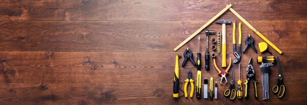 A variety of hand tools arranged on a wooden surface, forming the outline of a house, including pliers, screwdrivers, hammers, and rulers.