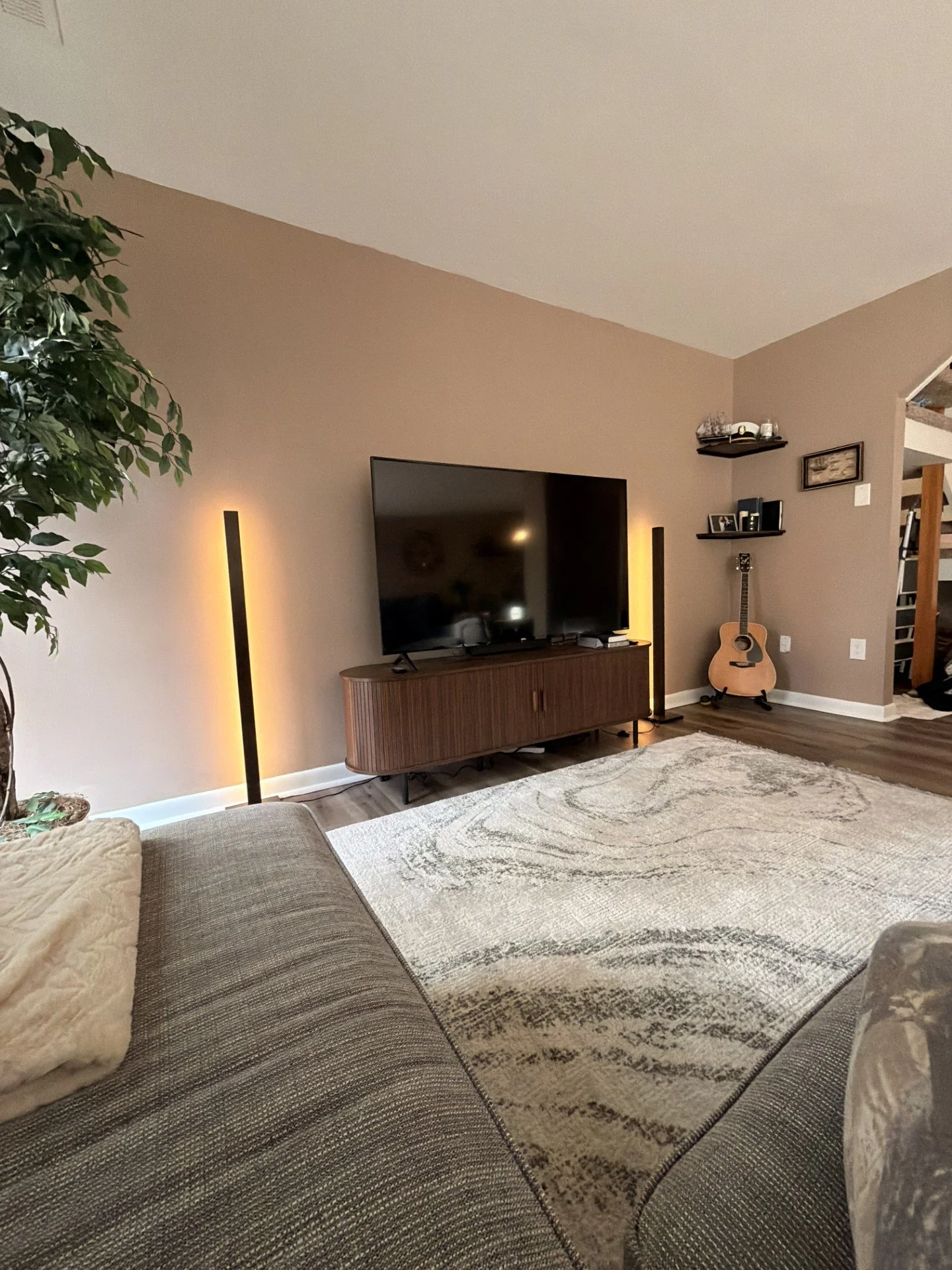 Living room with a large TV on a wooden console, illuminated by two tall floor lamps, and a guitar leaning against the wall near a shelf with decorative items. A beige area rug covers part of the hardwood floor, and a couch with a throw pillow is in the foreground.