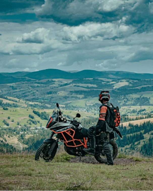 Person with helmet and backpack standing next to a motorcycle on a hilltop overlooking a green valley with rolling hills and a cloudy sky.
