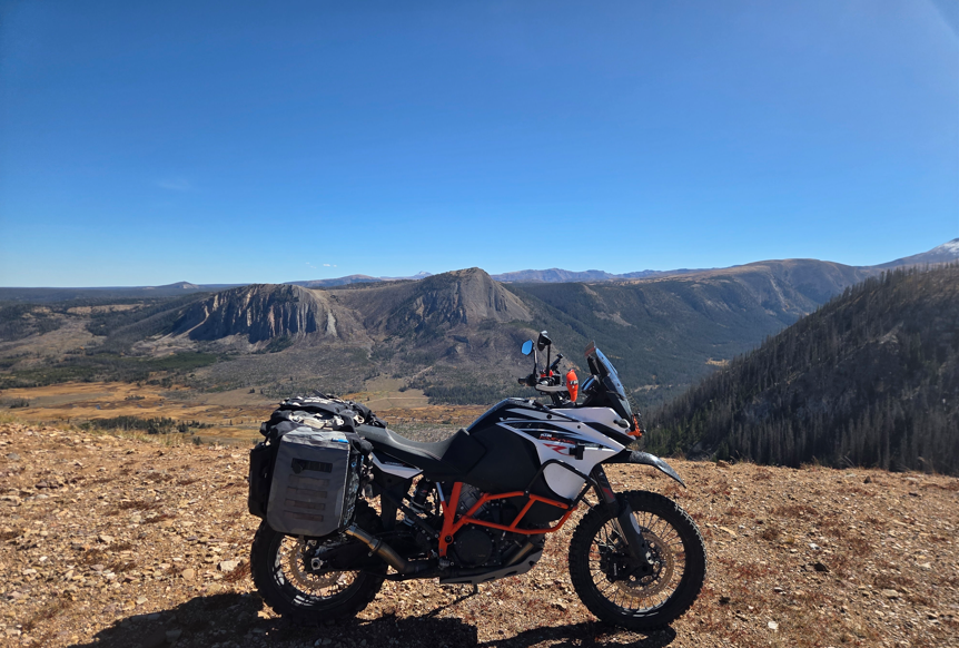 Motorcycle parked on a mountain overlook with expansive mountain range and clear blue sky in the background.