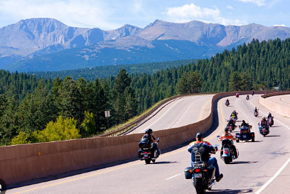Motorcycles riding on a curved mountain highway with trees and mountains in the background.