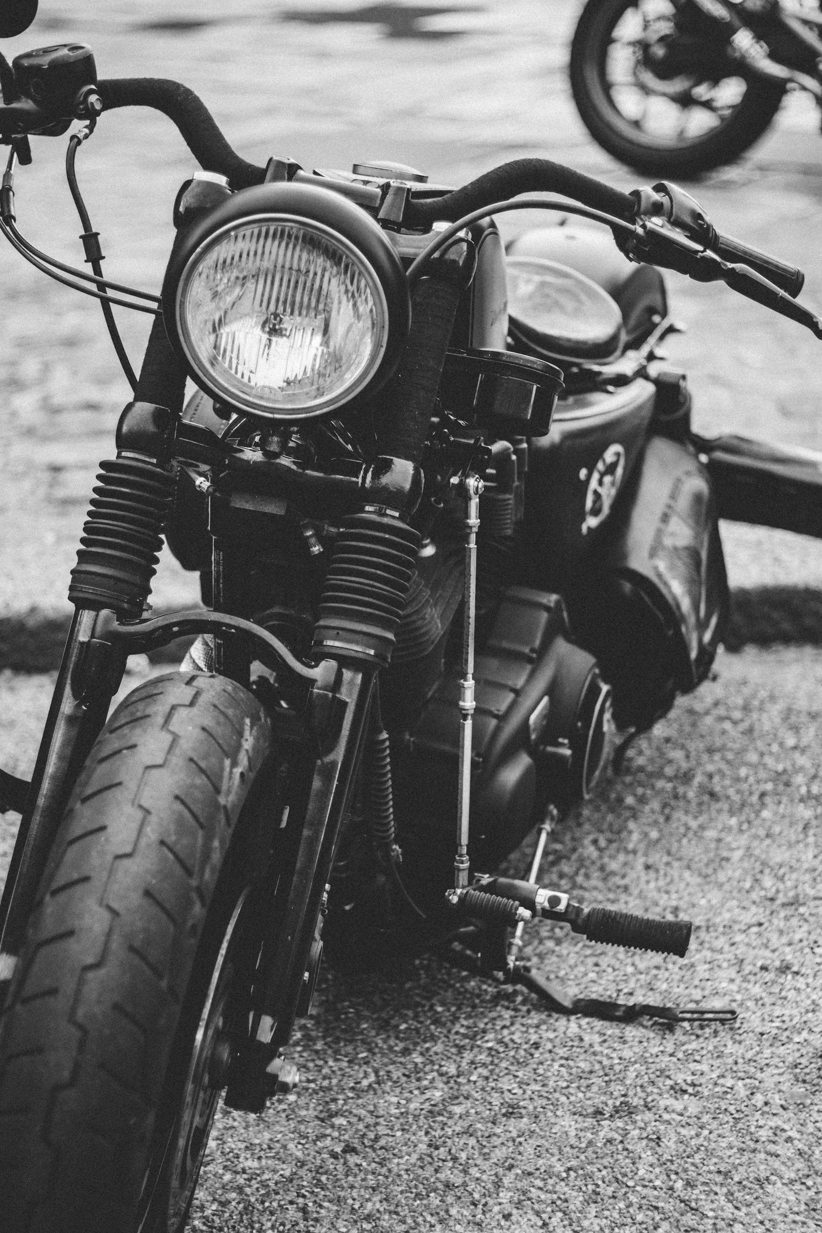 Black and white photo of a motorcycle with a large front tire, round headlight, and handlebars, parked on a textured ground surface, with another motorcycle partially visible in the background.