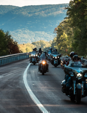 Group of motorcyclists riding on a curvy mountain road surrounded by trees and mountains in the background.