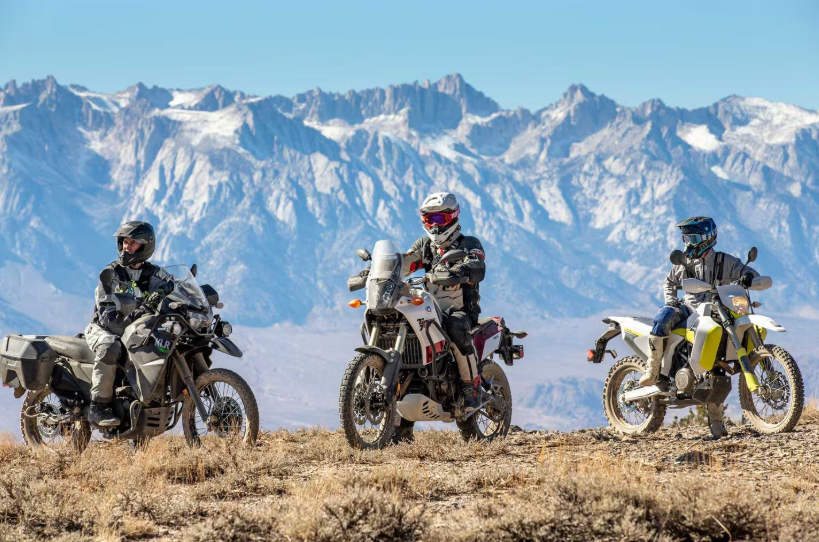 Three motorcyclists in full gear riding adventure bikes on a dry, grassy terrain with snow-capped mountains in the background.