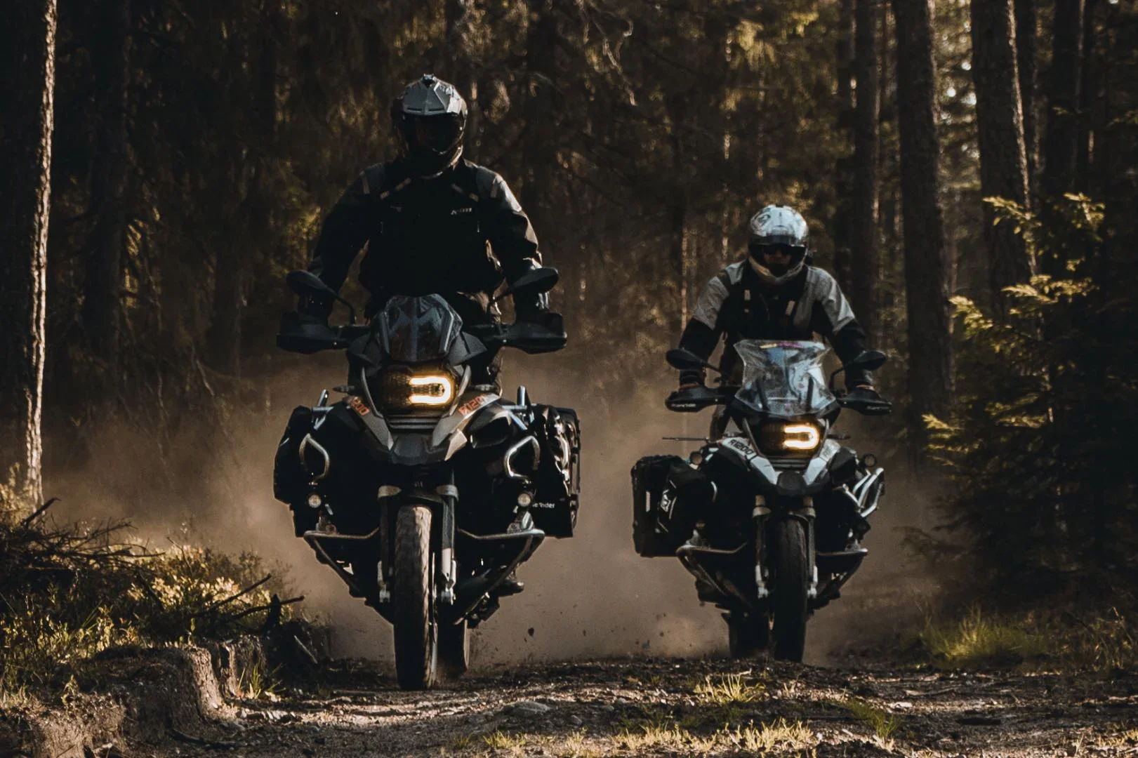 Two motorcycle riders wearing helmets riding through a forest trail, kicking up dust, during what appears to be late afternoon or early evening.