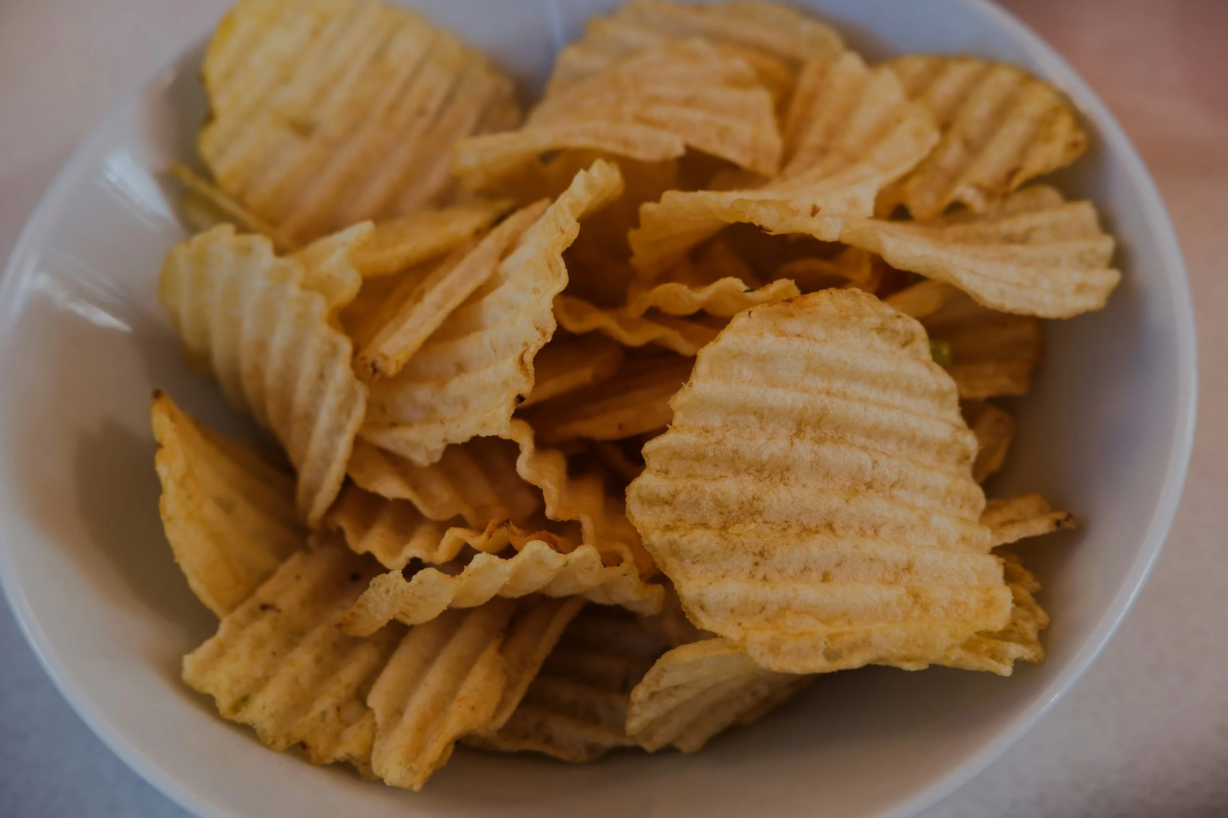 A white bowl filled with ridged potato chips on a light-colored surface.