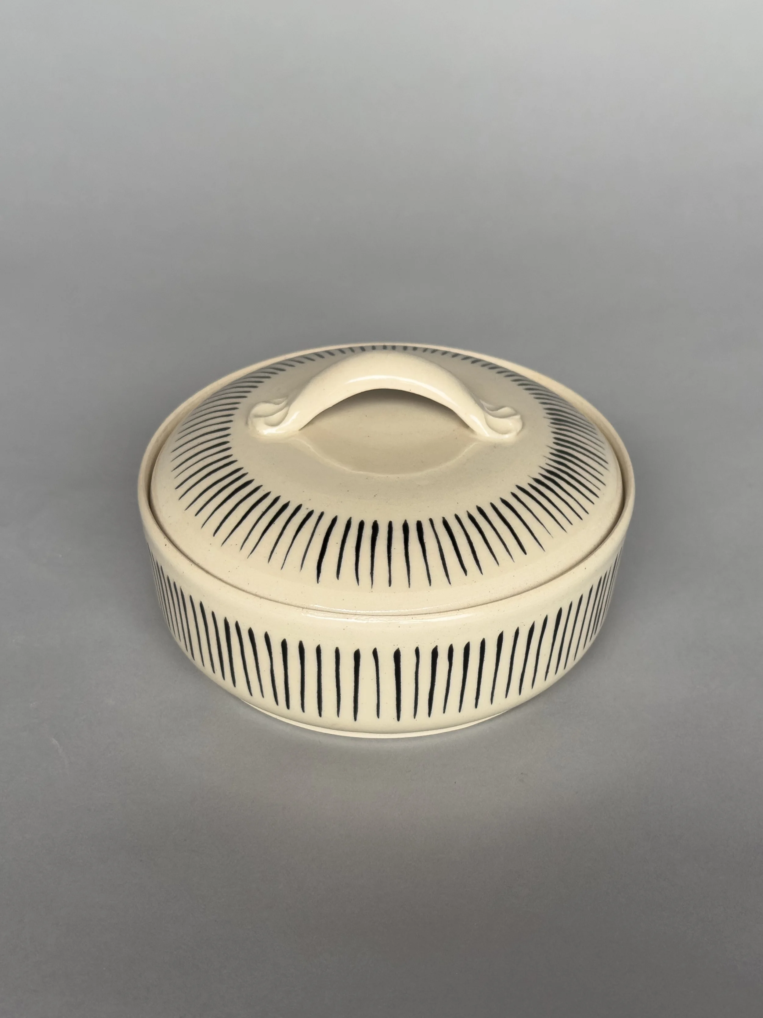 Handmade ceramic covered dish with black line pattern and lid handle by Jeanette Burgett Ceramics on a gray background.