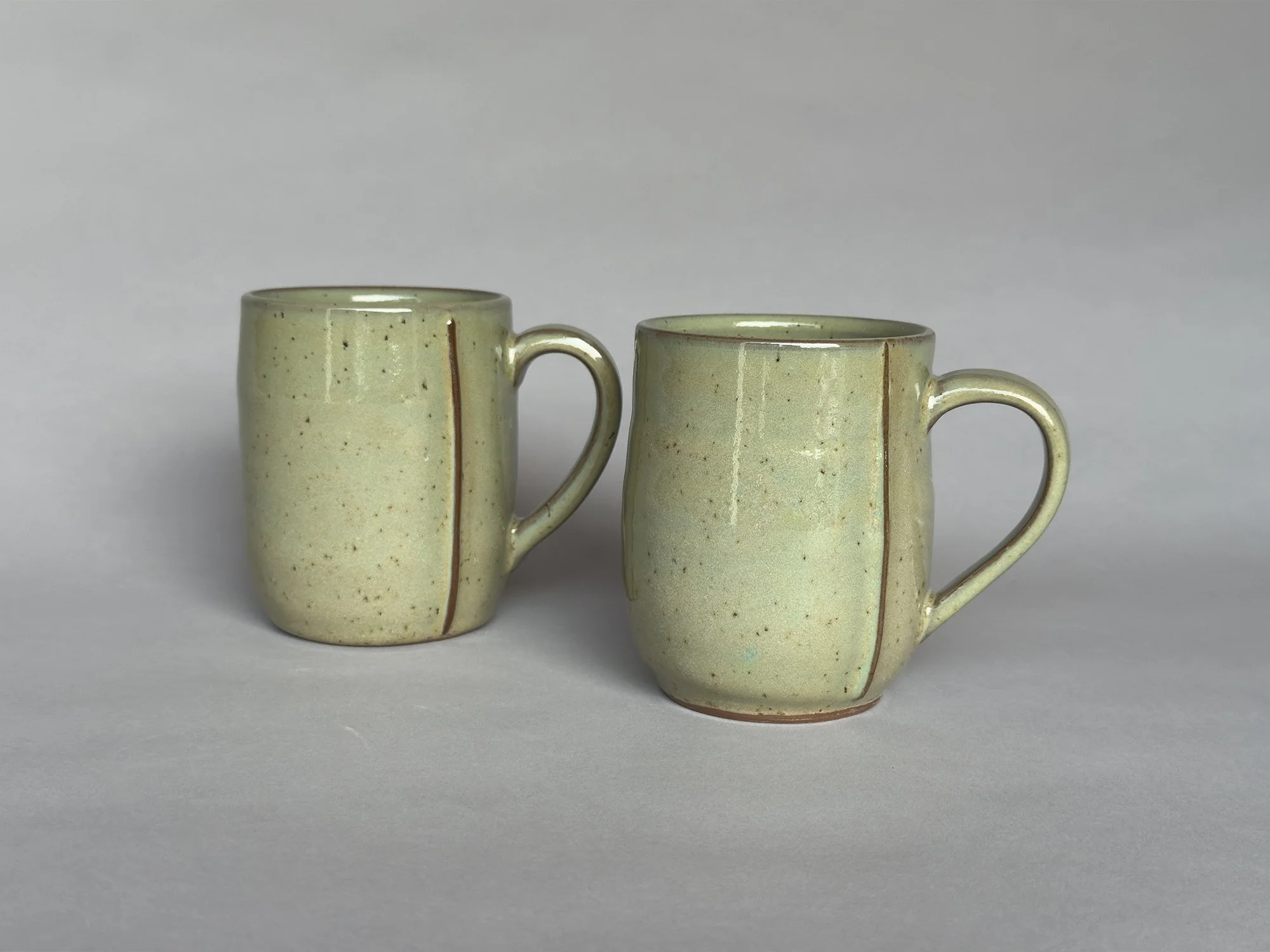 Two handmade ceramic coffee mugs with speckled sage green glaze and curved handles by Jeanette Burgett Ceramics, photographed on a soft gray background.