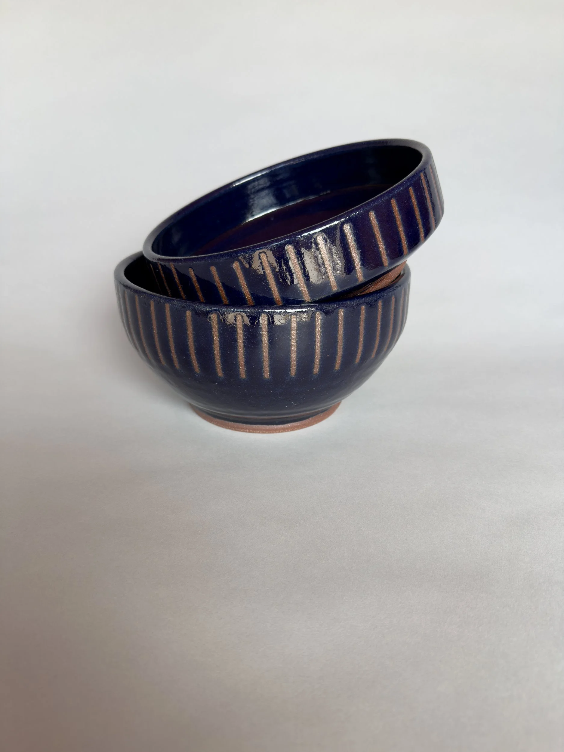 Two handmade deep blue ceramic bowls with carved vertical stripe details and glossy glaze by Jeanette Burgett Ceramics on a soft gray background.