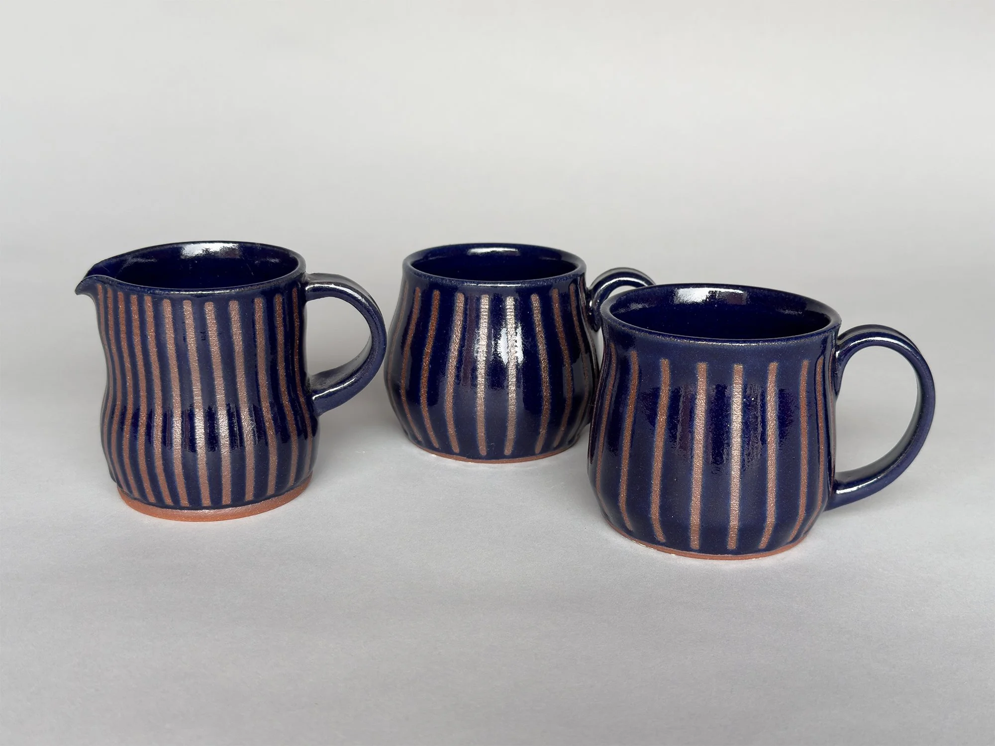 Handmade deep blue ceramic creamer pitcher and matching mugs with carved vertical stripe details and glossy glaze by Jeanette Burgett Ceramics on a soft gray background.