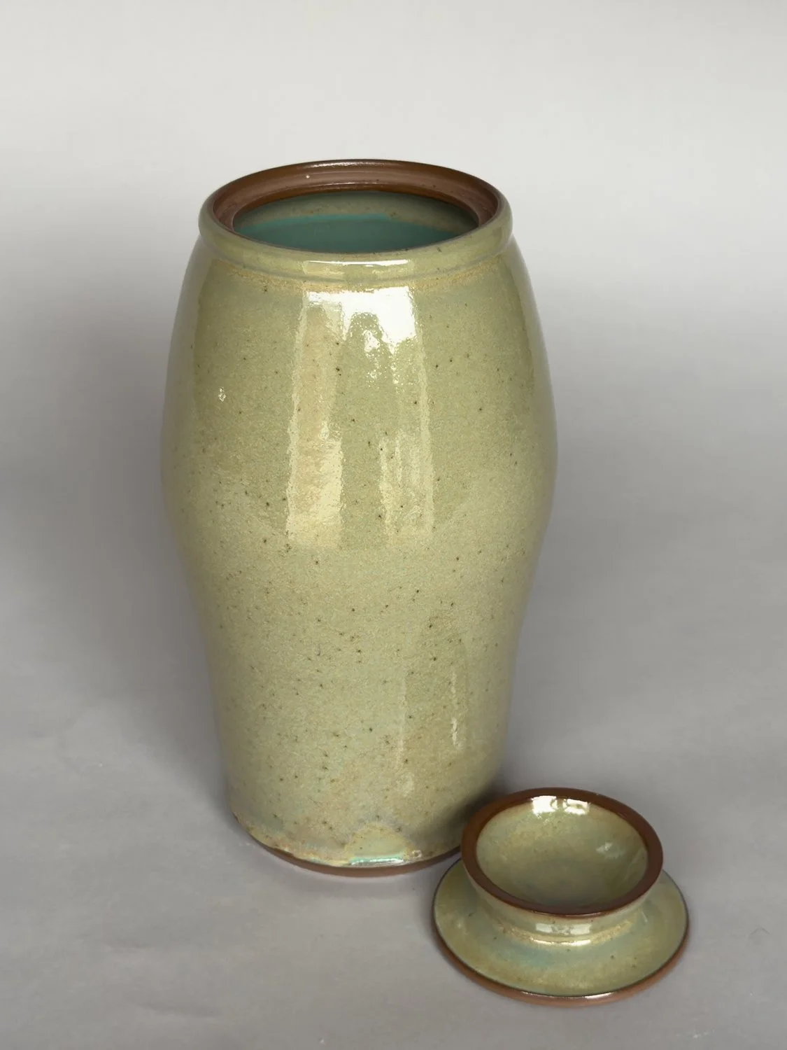 Tall handmade ceramic vase with lid and soft speckled sage green glaze by Jeanette Burgett Ceramics on a neutral gray background.