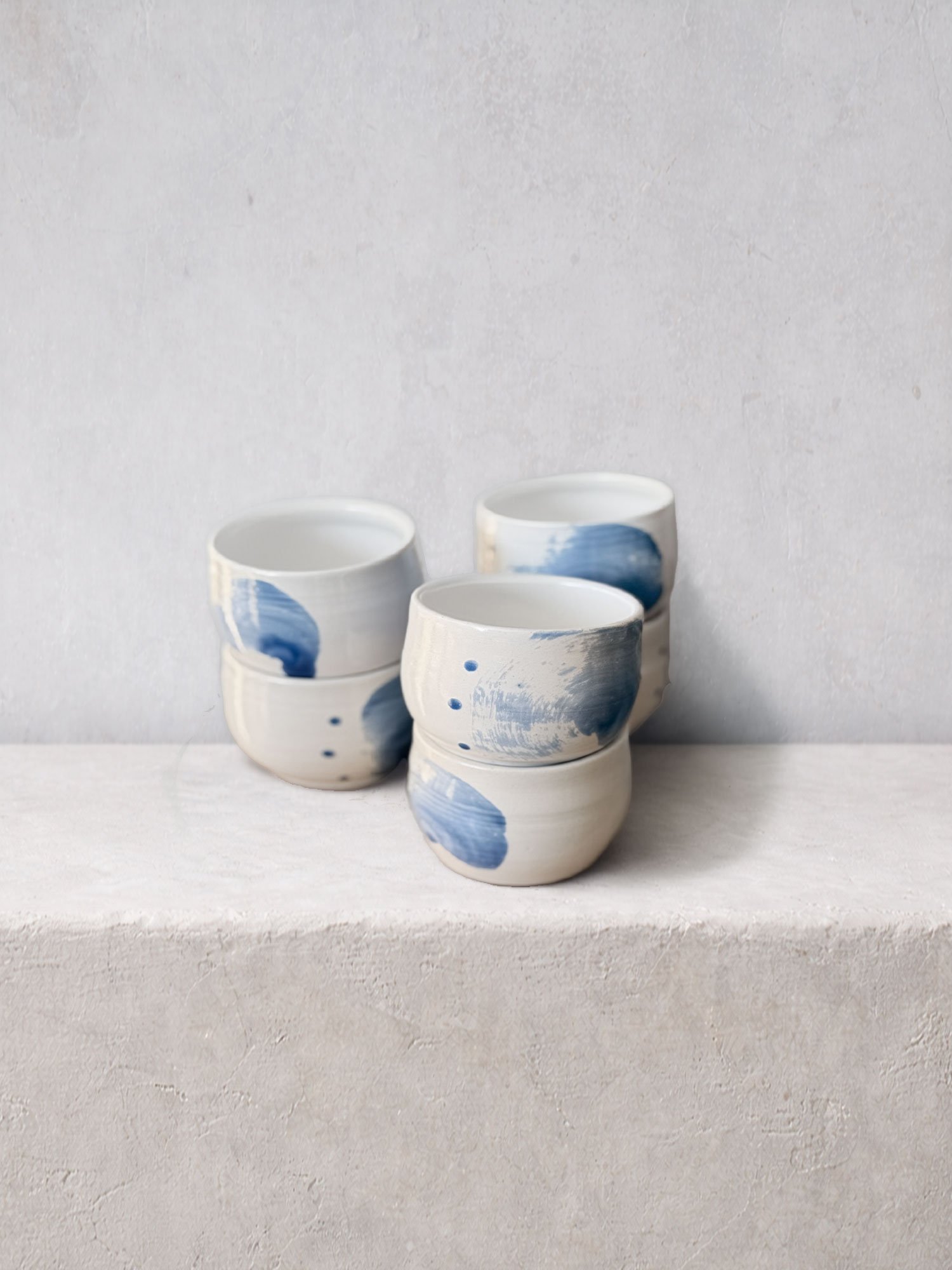 Set of handmade ceramic cups featuring soft white clay and expressive blue brushwork, arranged on a textured surface.
