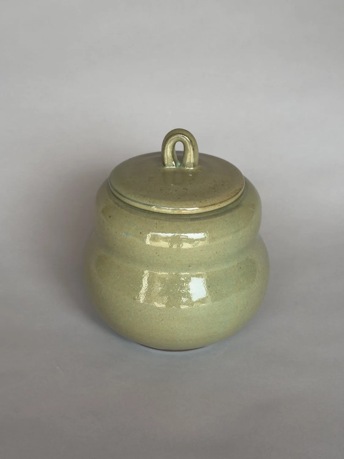 Rounded handmade ceramic lidded jar with glossy sage green glaze by Jeanette Burgett Ceramics on a neutral gray background.