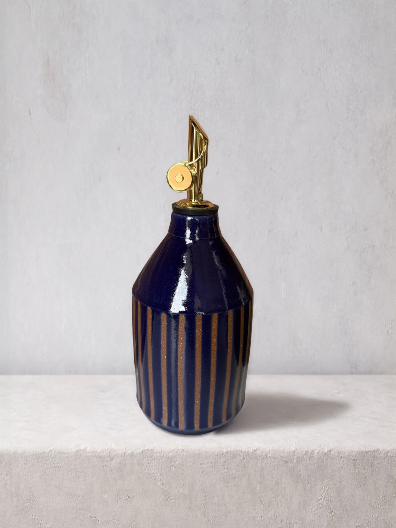 Handmade ceramic oil bottle with deep blue glaze, vertical texture, and a brass pour spout, photographed on a minimalist studio surface.