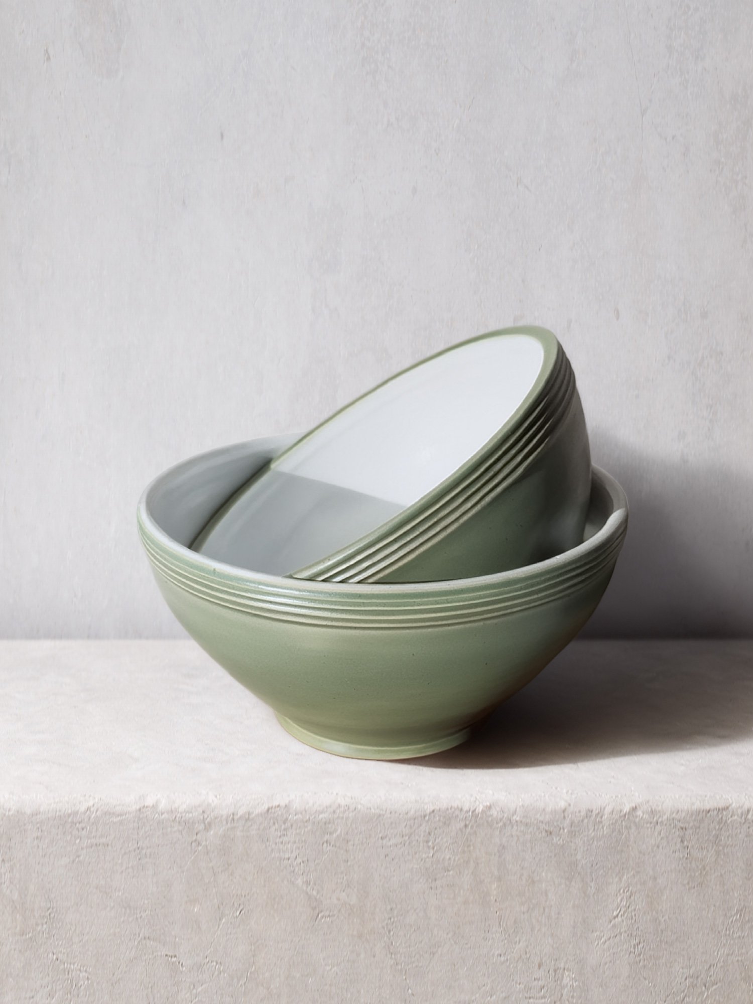 Stacked handmade ceramic bowls with a soft green glaze and subtle ridged detail, photographed on a textured plaster surface.